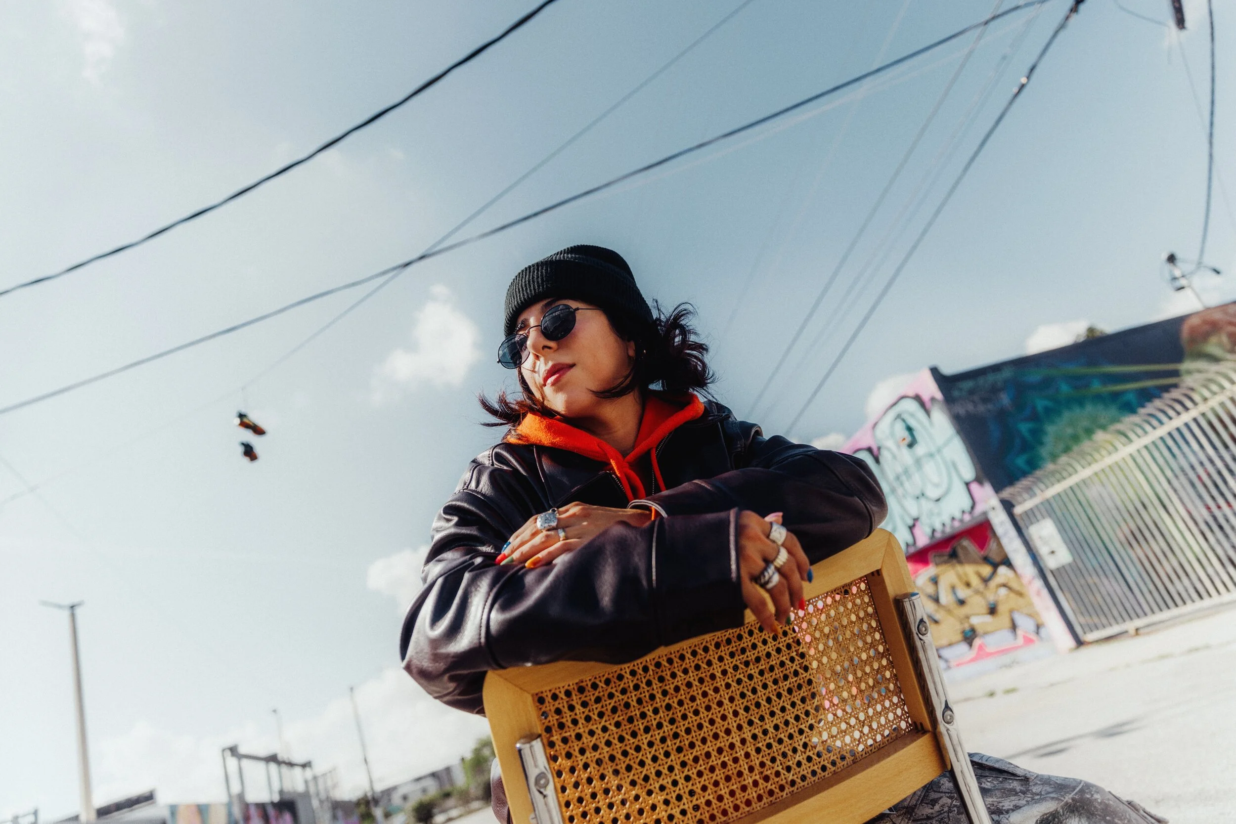 A woman wearing sunglasses, a black beanie, and a leather jacket with an orange hoodie, sitting on a chair outdoors with colorful graffiti in the background.