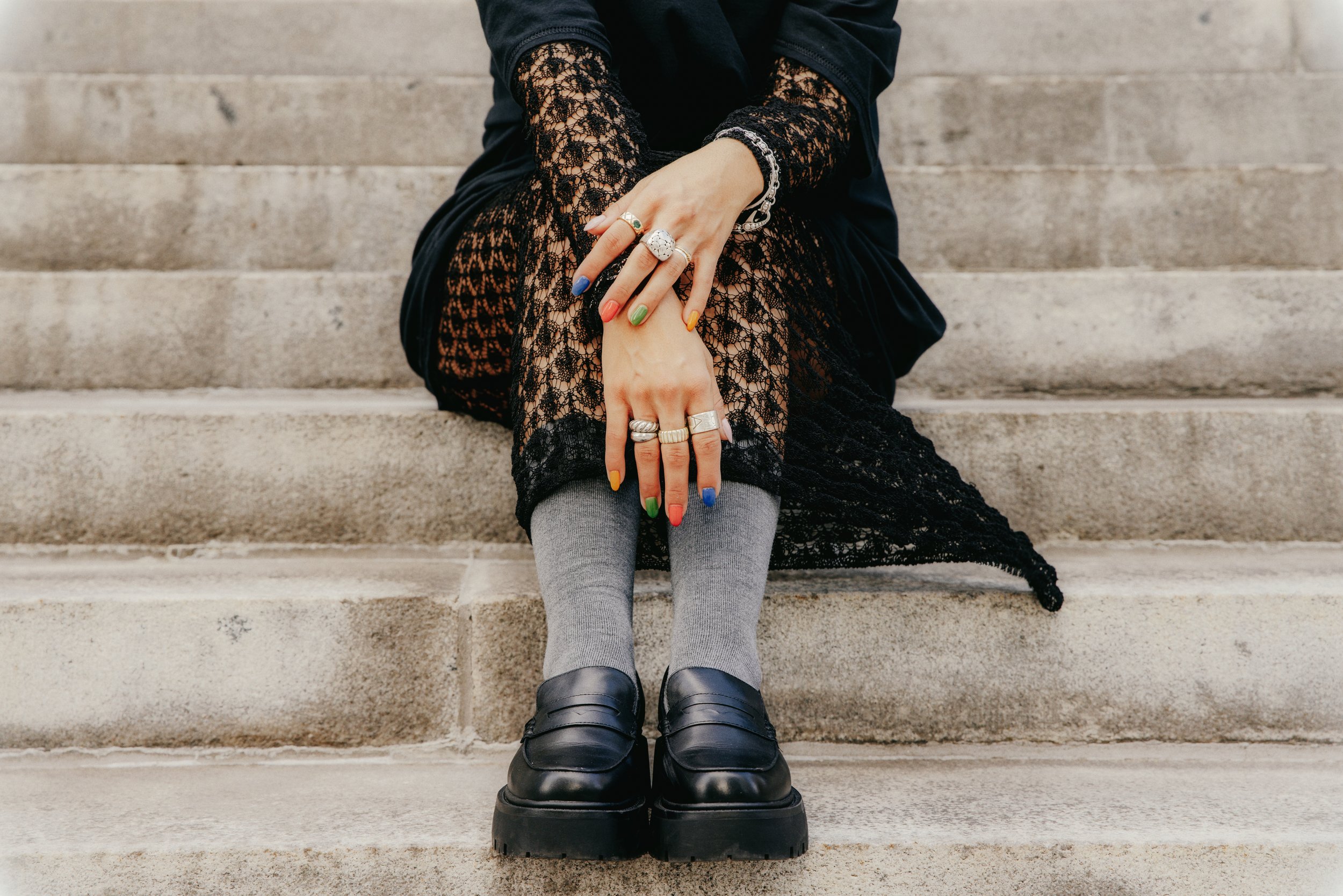 Person sitting on stone steps, wearing black lace dress, gray knee-high socks, and black loafers, with colorful fingernails and multiple rings on fingers.