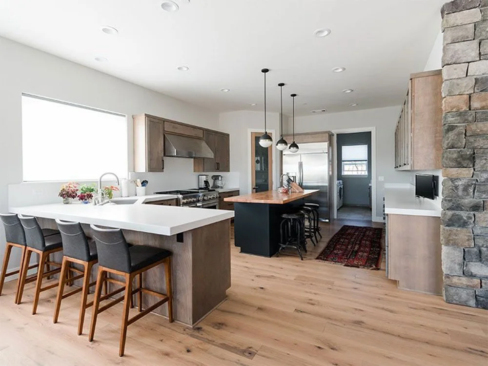 Modern kitchen with white countertops, wooden cabinets, black island with pendant lights, stainless steel appliances, and a stone accent wall.