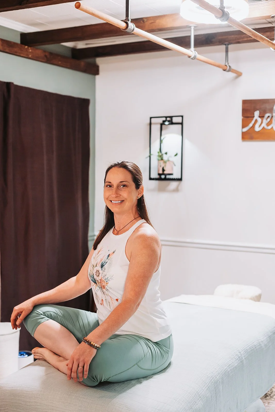 Jessi sitting cross-legged on her massage table, smiling and exuding confidence.