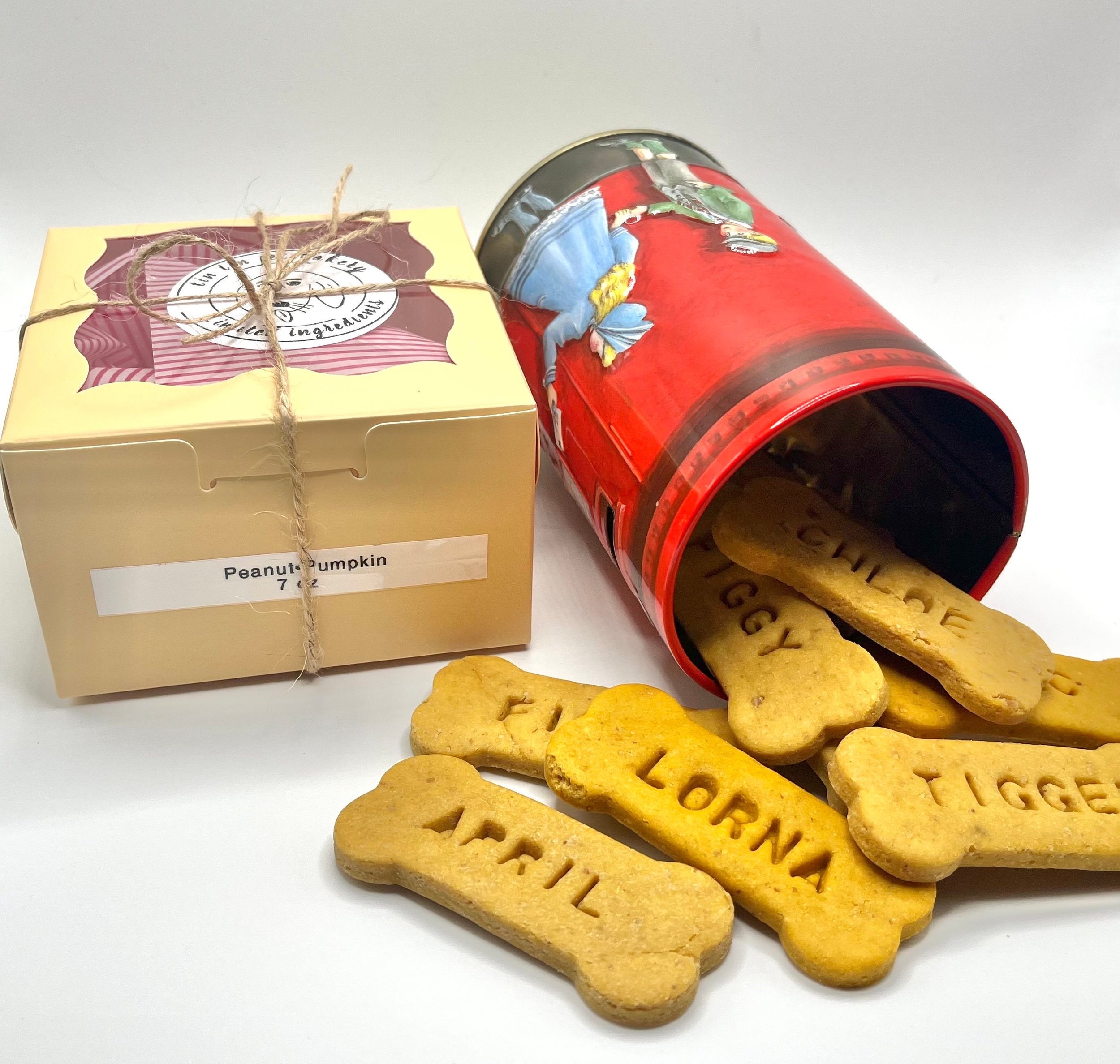 A box of peanut pumpkin cookies labeled 'Peanut Pumpkin 7 oz', a red tin container tipped over with 'LORNA' and 'TIGER' embossed cookies spilling out, some stamped with 'TRI'. The cookies are bone-shaped and placed on a plain white background.