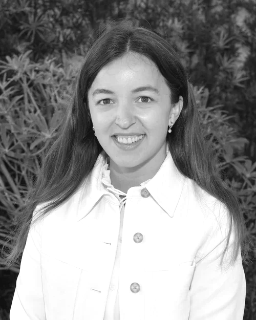 A smiling young woman with long hair, wearing earrings and a white jacket, standing outdoors in front of leafy plants.