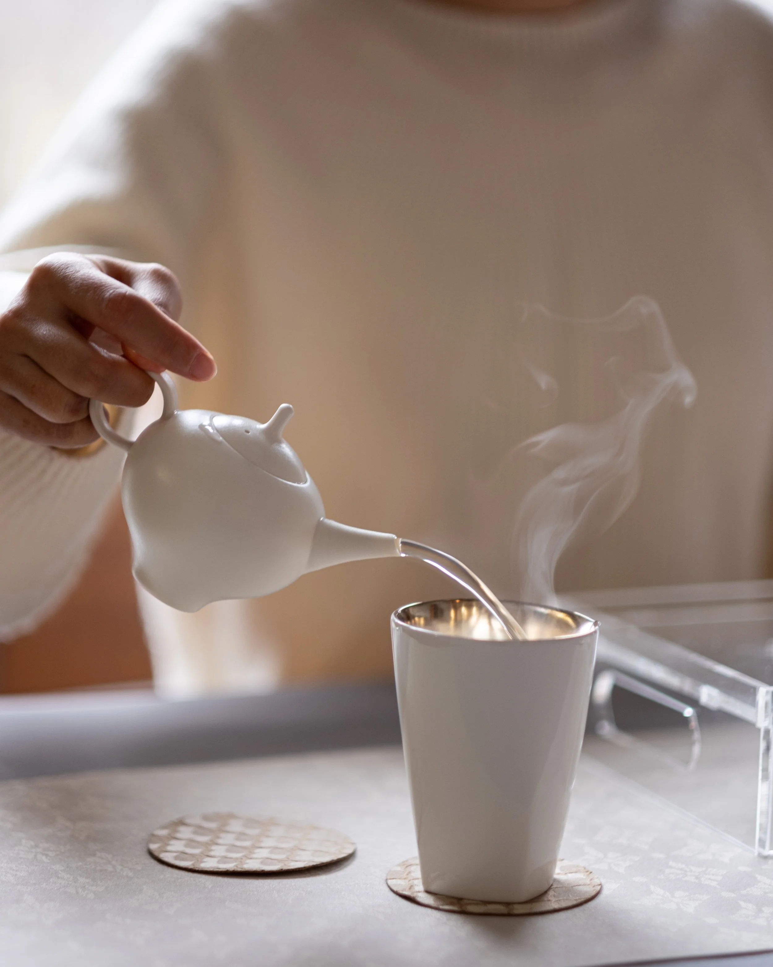 Hot water is poured from a white teapot into a cup, with steam rising.