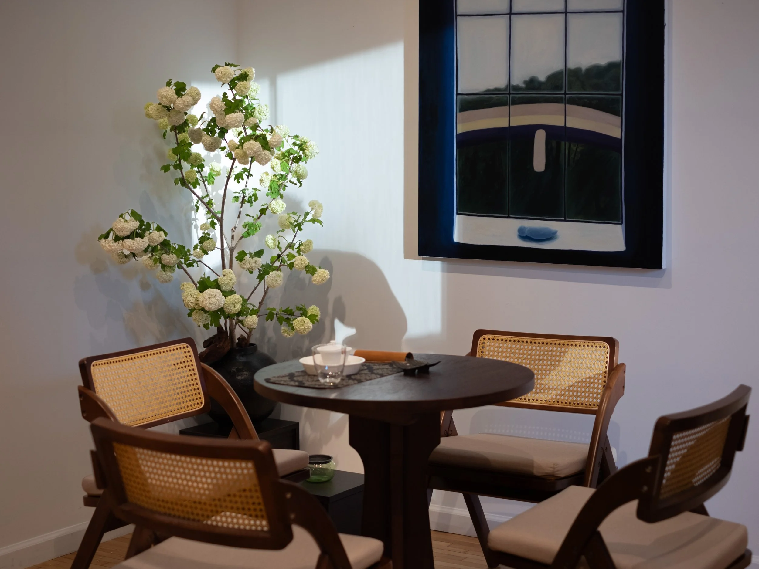 Interior of a dining room with a round wooden table and four wooden chairs with woven backs. A large vase with white hydrangea flowers is in the corner, and a framed painting of a window with a view of nature hangs on the wall.