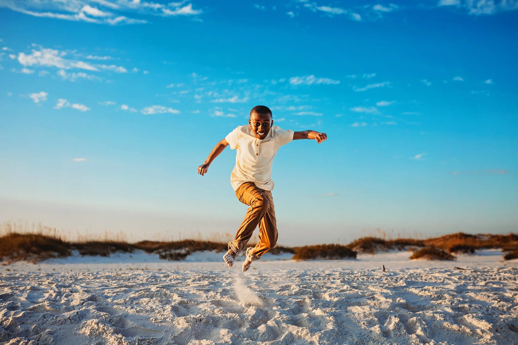 A young boy jumps off of a sand cliff at Pensacola Beach with a beautiful blue sky behind him. Taken by Pensacola Photographer June Sunrise Photography.