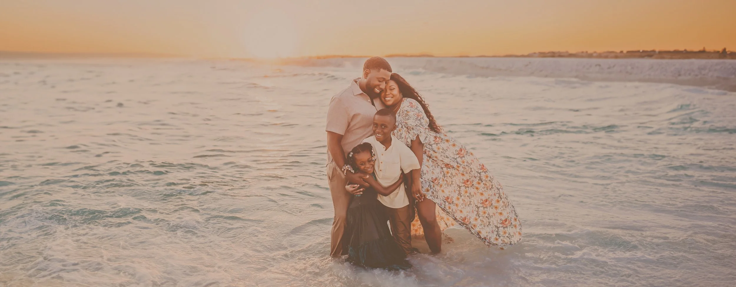 A family cuddles up close in front of a beautiful sunset on Pensacola Beach, FL while mom wears a flower patterned dress and the glow of the sunset illuminates their skin. Taken by Pensacola Photographer June Sunrise Photography.