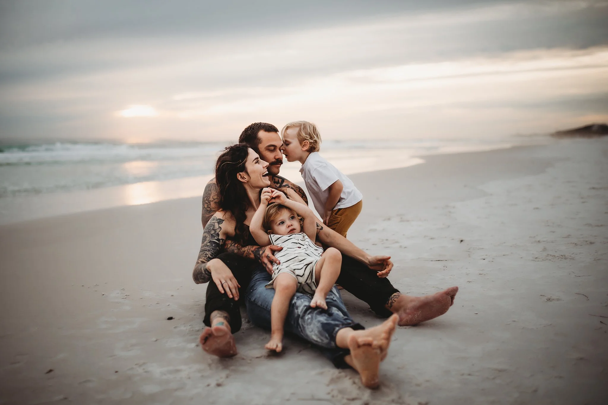A family sits on the beach near the shore line while their youngest son sits in the mother's lap and the older son whispers something in his father's ear. Taken by Pace FL family Photographer June Sunrise Photography.