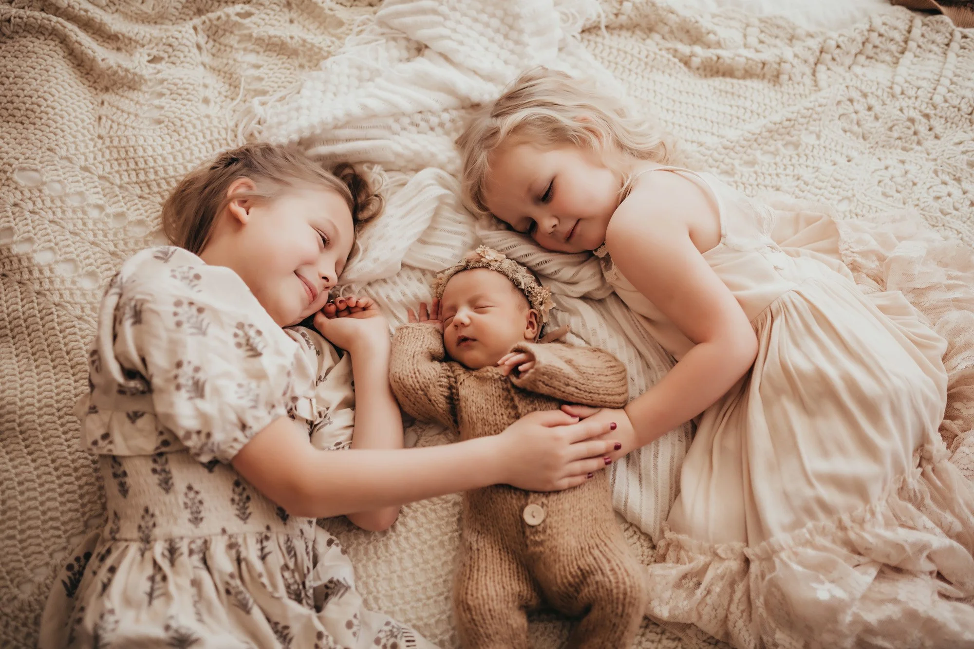 Two older sisters smile and snuggle their newborn baby sister as they all are wearing neutral beige tones and laying on a bed of layered beige and white blankets.