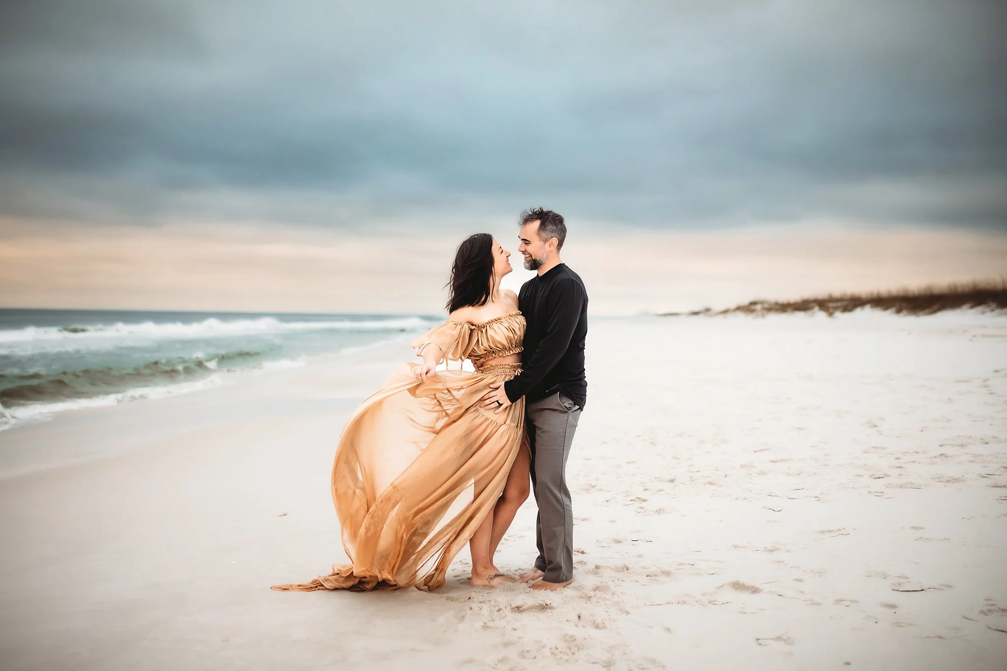 A husband and wife cuddle on the beach in Destin, Florida as the woman's beige dress blows in the wind. Taken by Destin Photographer June Sunrise Photography.