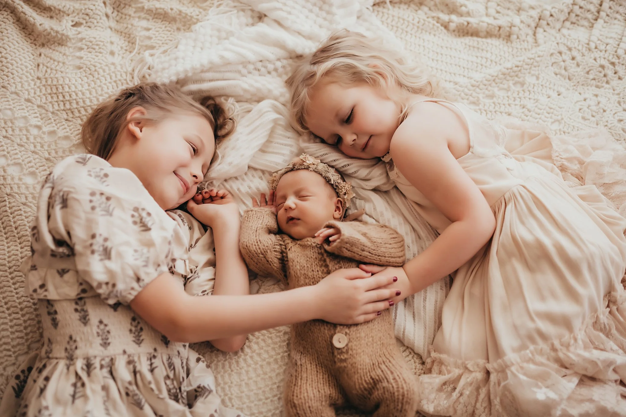 Two big sisters snuggle with their newborn baby sister in their home while all dressed in neutrals and laying on a cream colored crotchet blanket. Taken by Pace Florida Newborn Photographer June Sunrise Photography.