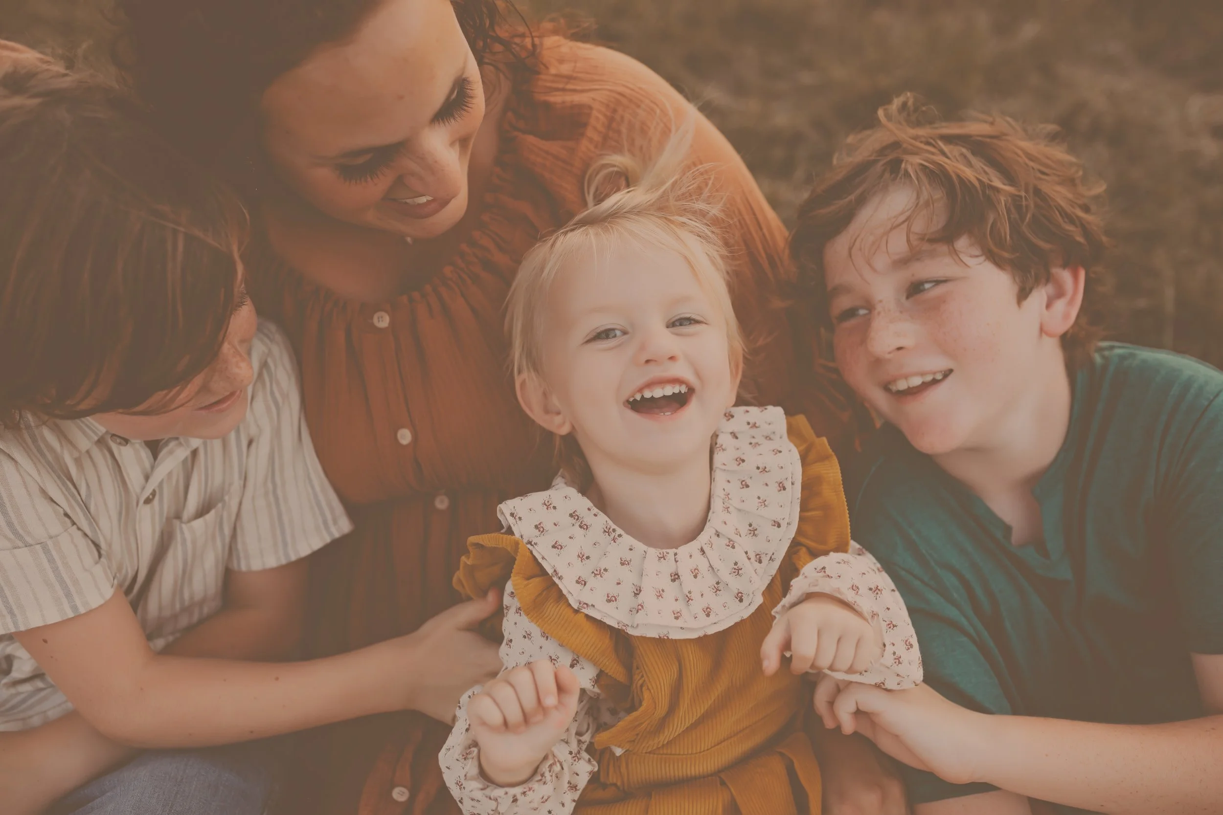 A mom snuggles up with her three children while they sit next to her and on her lap and everyone smiles and laughs while interacting with each other.Taken by Pensacola Photographer June Sunrise Photography.