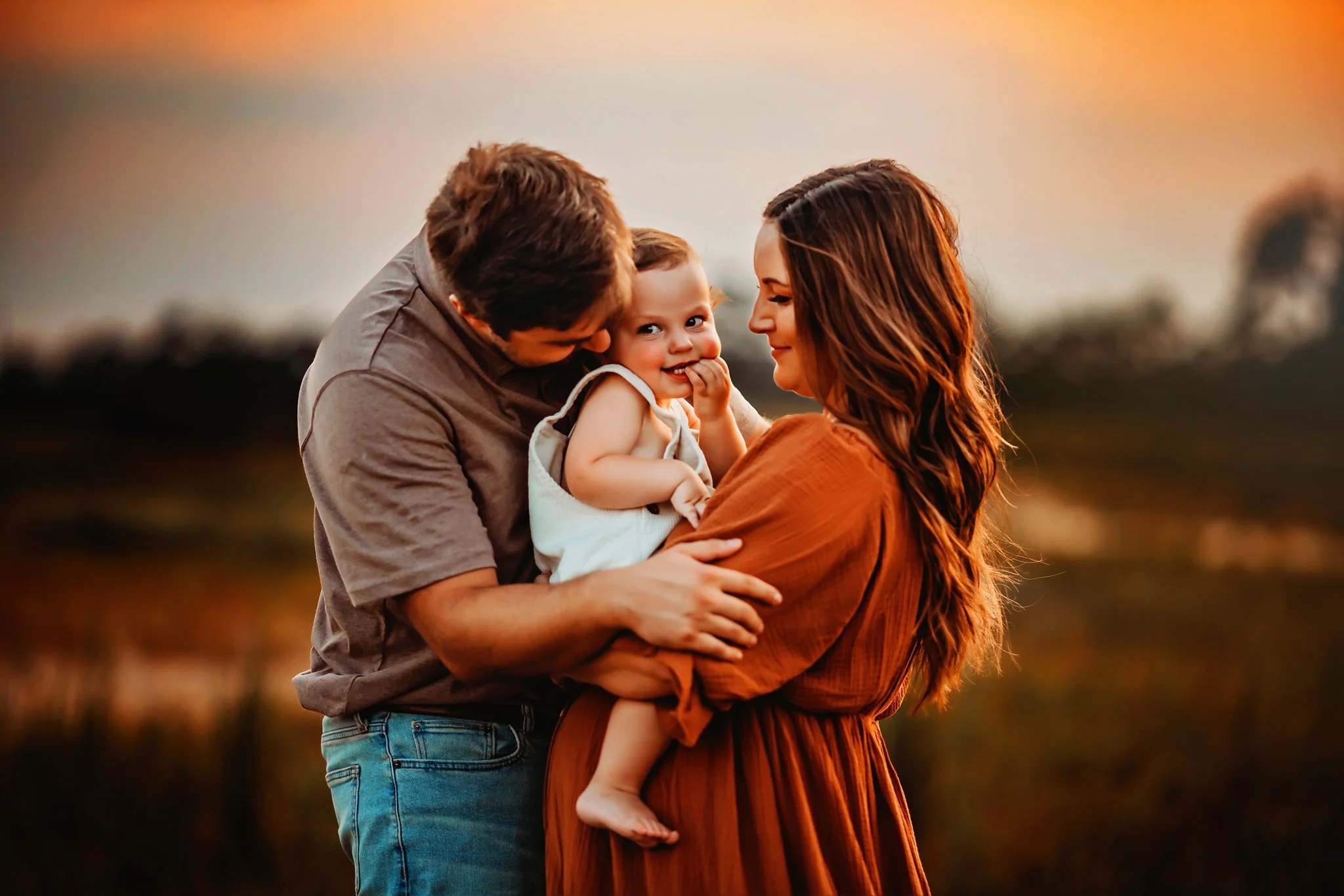 A mother and father snuggle their toddler son close during their sunset family maternity session in Pace, Florida. Taken by Pace FL family Photographer June Sunrise Photography.