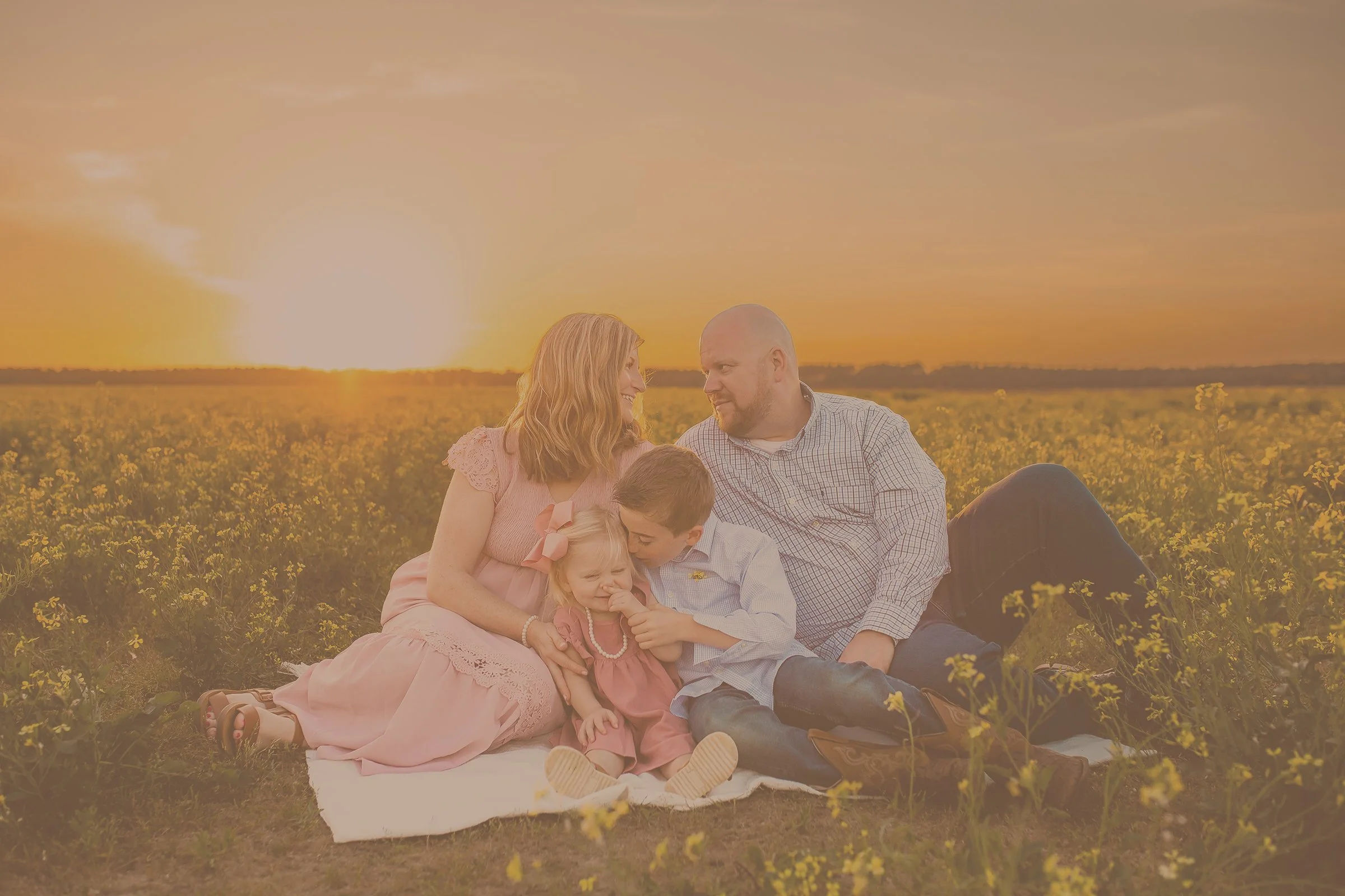 A mom and dad look lovingly at each other while their children in their laps smile and whisper secrets to each other. Taken by Pensacola Photographer June Sunrise Photography.