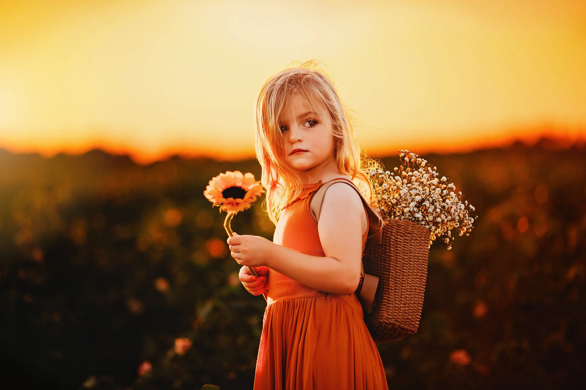 A toddler girl looks off into the distance while standing in front of a beautiful sunset and wearing a backpack full of baby's breath flowers in Pace, Florida. Taken by Pace FL family Photographer June Sunrise Photography.