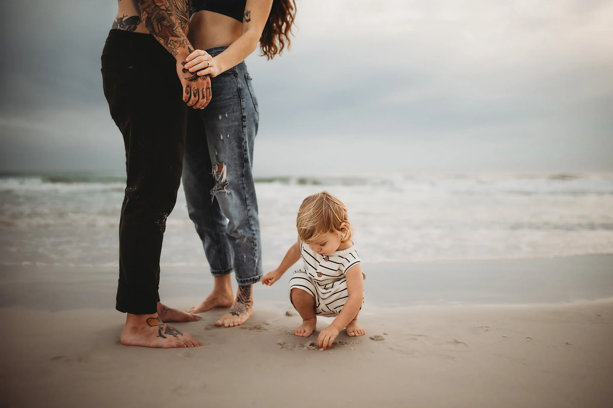 A toddler boy plays in the sand at his mother and father's feet on the beach in Destin, Florida. Taken by Destin Photographer June Sunrise Photography.