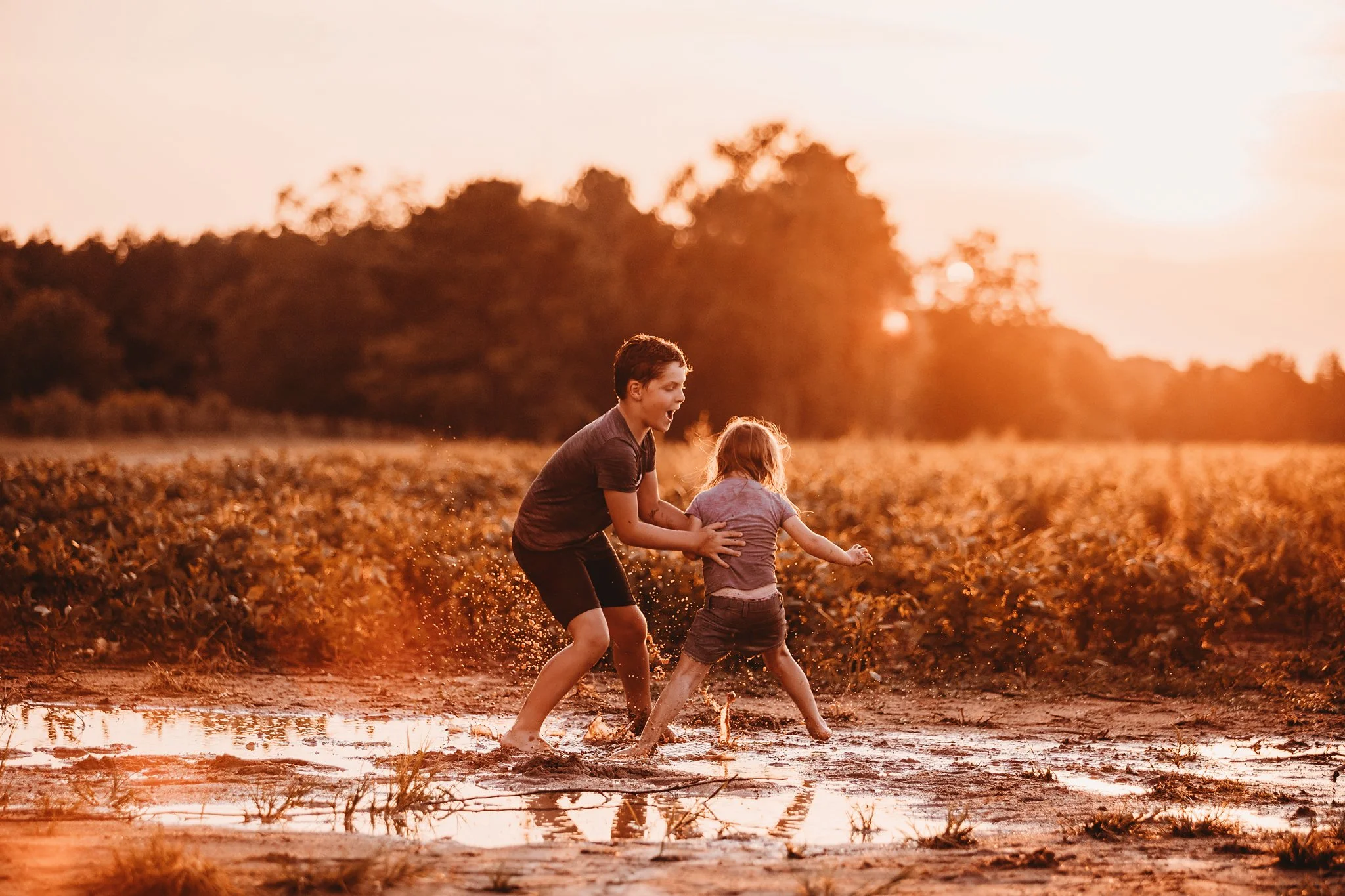 A young brother and sister play in a mud puddle in front of a beautiful sunset in Pace, Florida. Taken by Pace FL family Photographer June Sunrise Photography.
