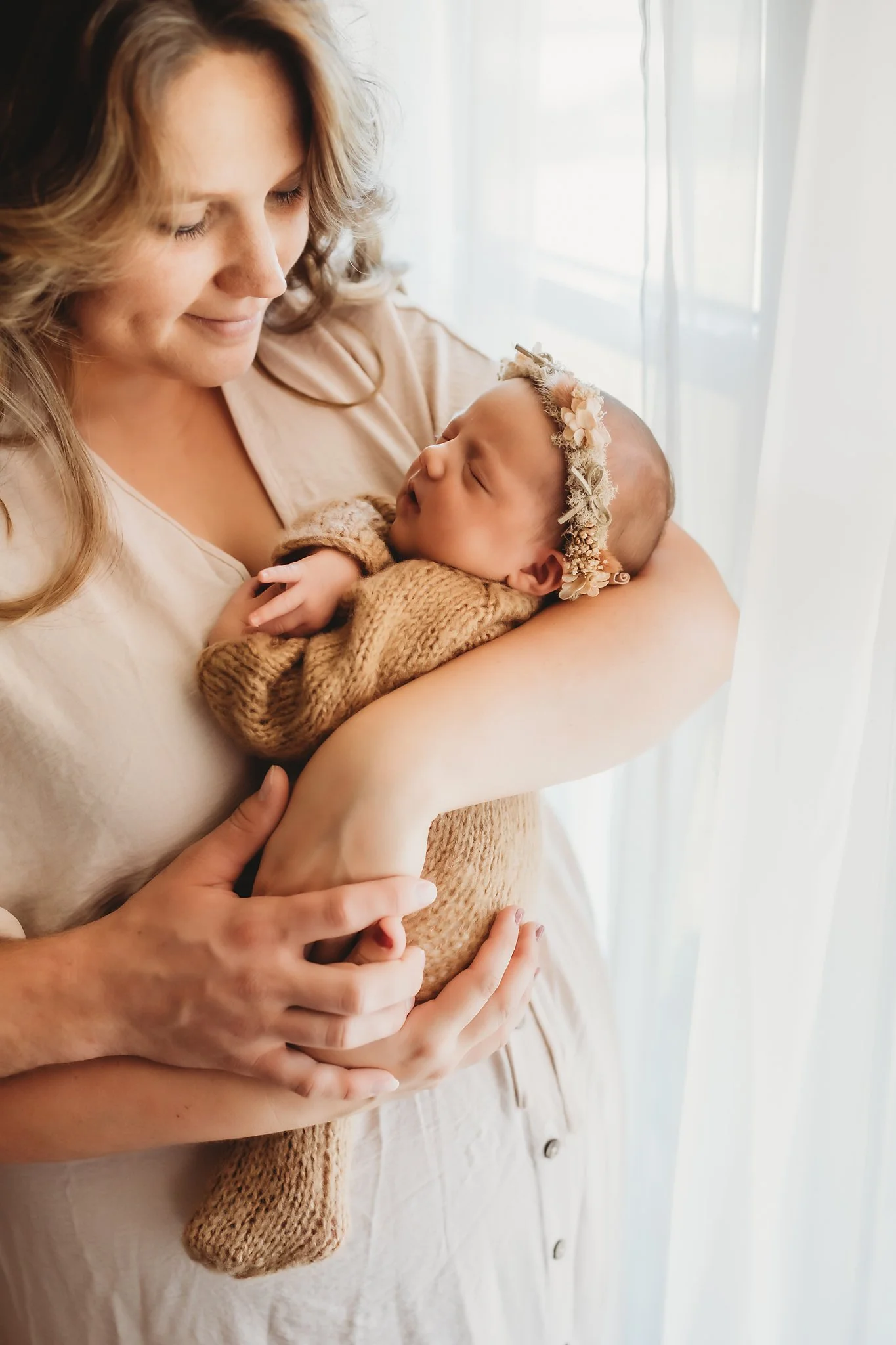A mother looks down at her newborn baby girl in her arms and smiles in front of a window in her home. Taken by Pace FL newborn Photographer June Sunrise Photography.