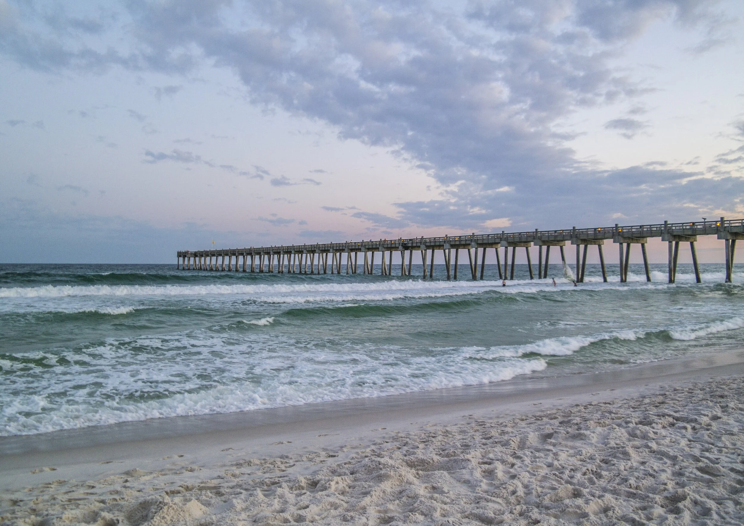 A sandy beach with gentle ocean waves and a wooden pier extending into the water, under a partly cloudy sky during dusk or dawn.