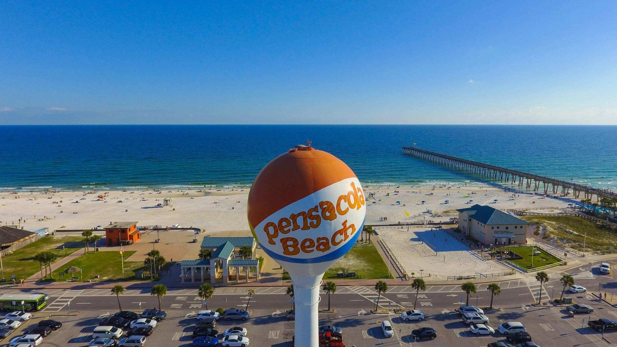A water tower with orange and white colors displays 'Pensacola Beach' near a sandy shoreline with people, a pier extending into the ocean, and a parking lot with cars and palm trees.