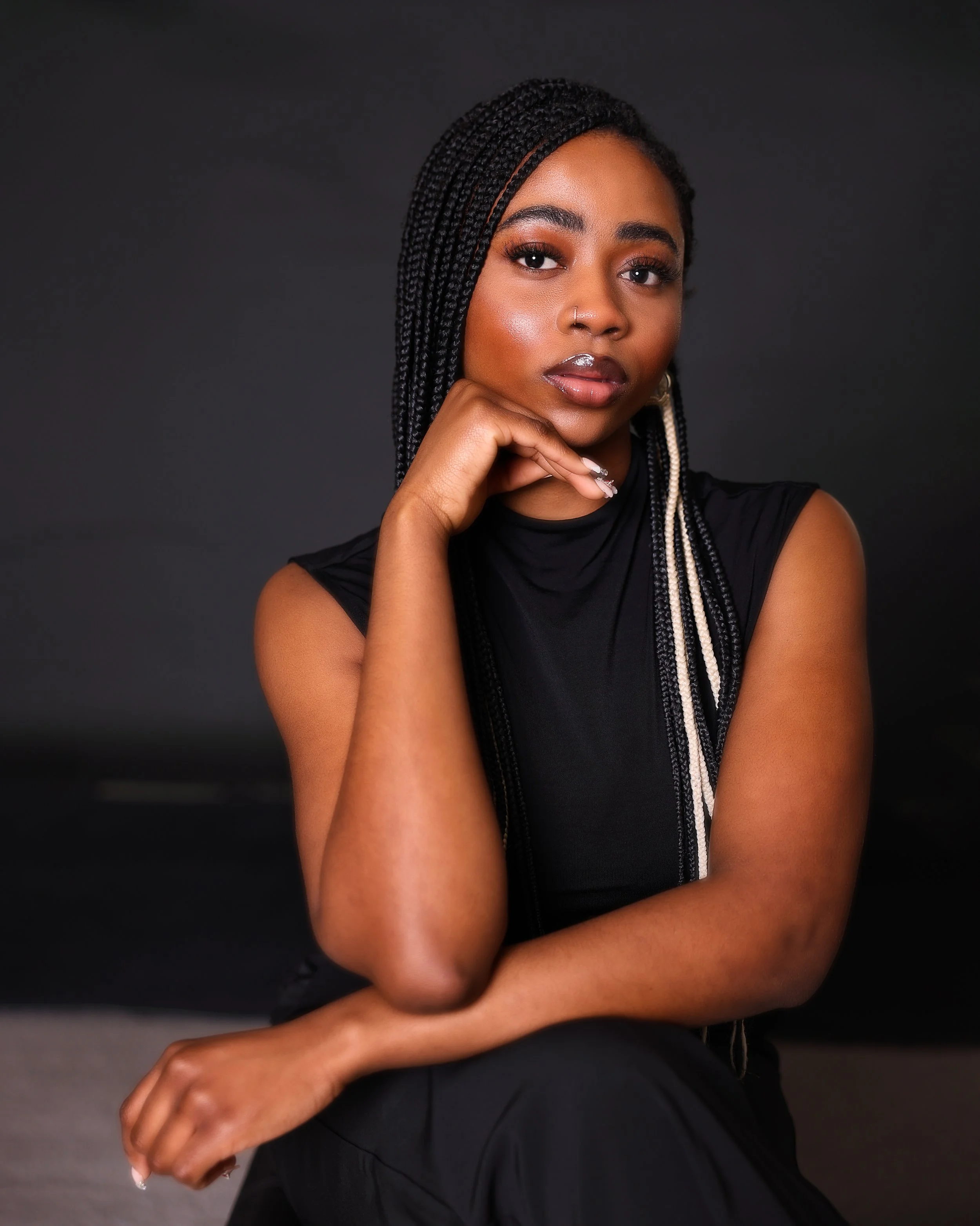 Portrait of a woman with long braided hair and makeup, wearing a sleeveless black top, seated against a dark background.