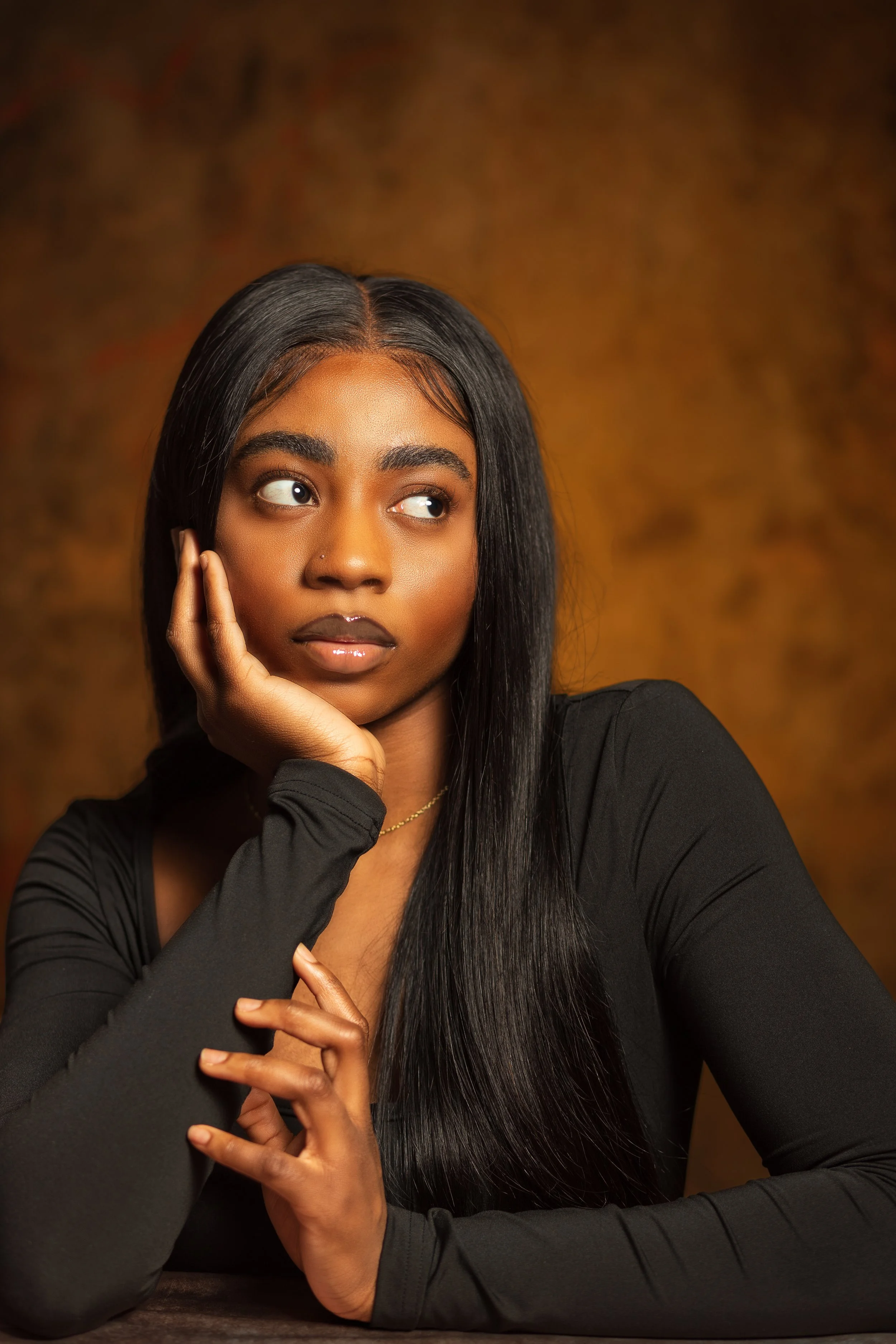 Portrait of a young African American woman with straight black hair, resting her face on her hand, wearing a black long-sleeve top, set against a warm, textured background.