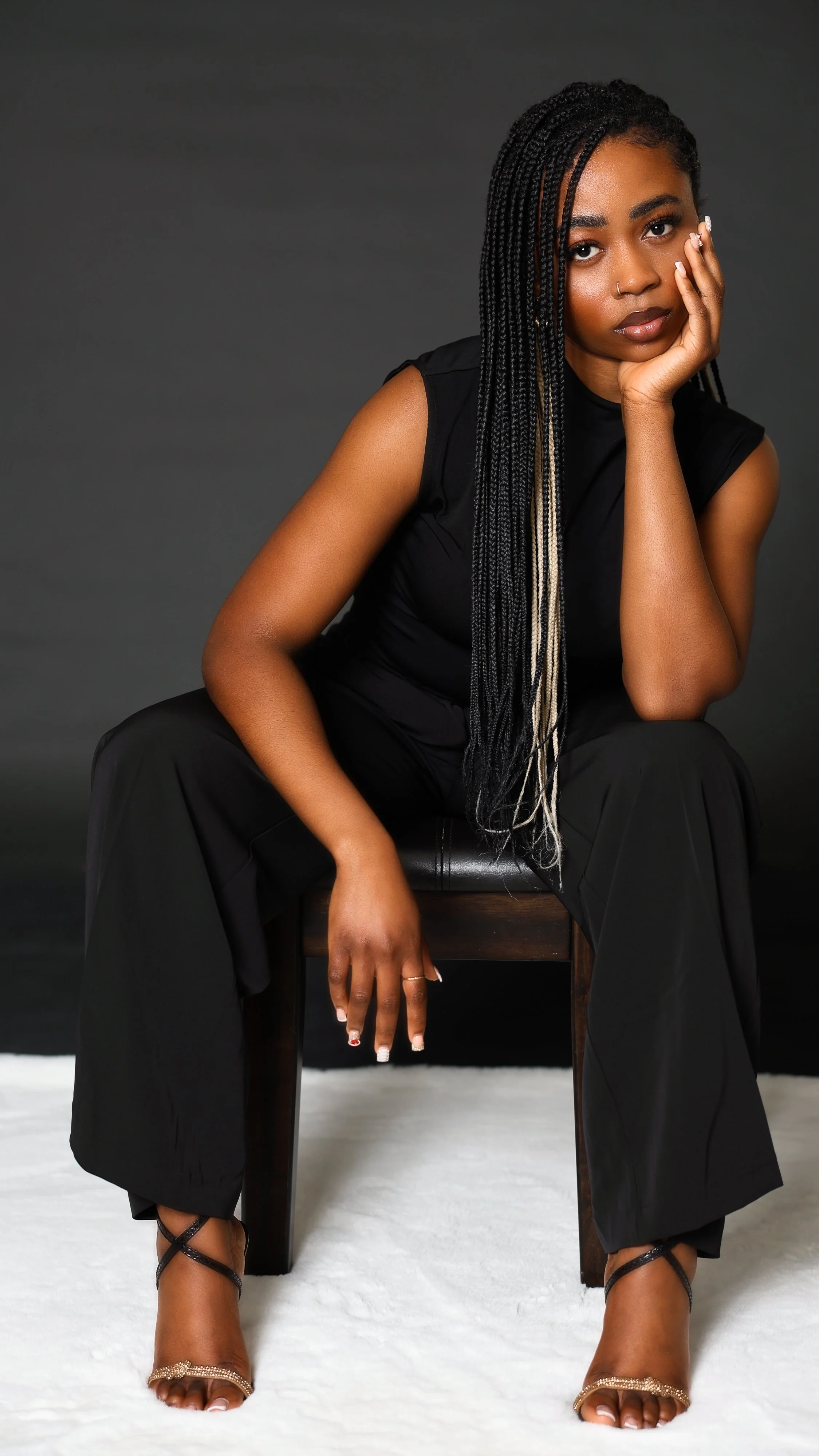 A woman with long braided hair and a nose ring sitting on a wooden bench against a black background, resting her chin on her hand, wearing black pants and a sleeveless top, with her legs spread apart and wearing strappy heels.