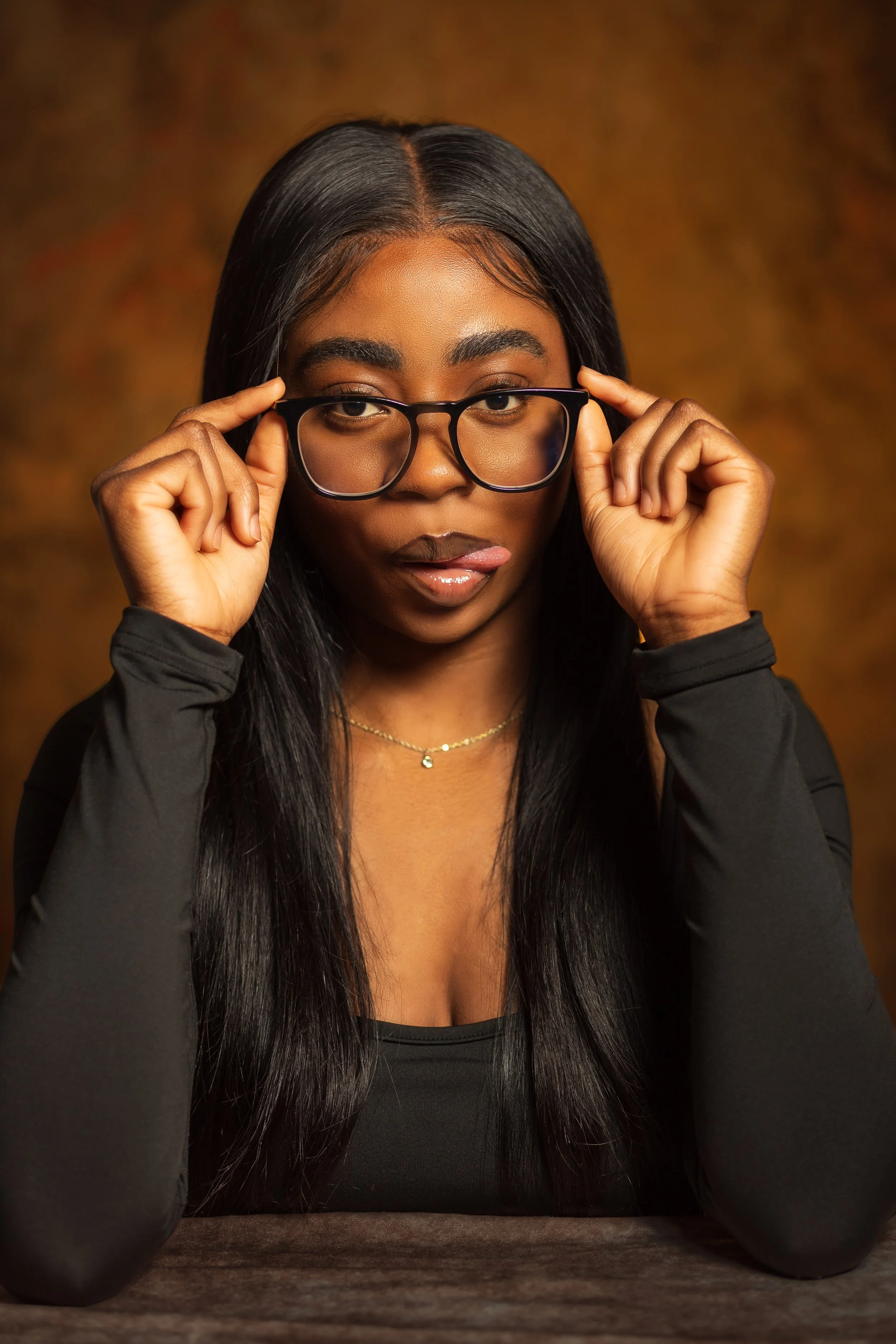 A young woman with long black hair, wearing glasses and a black long-sleeve top, looks into camera with a serious expression, holding her glasses with both hands, against a warm brown background.