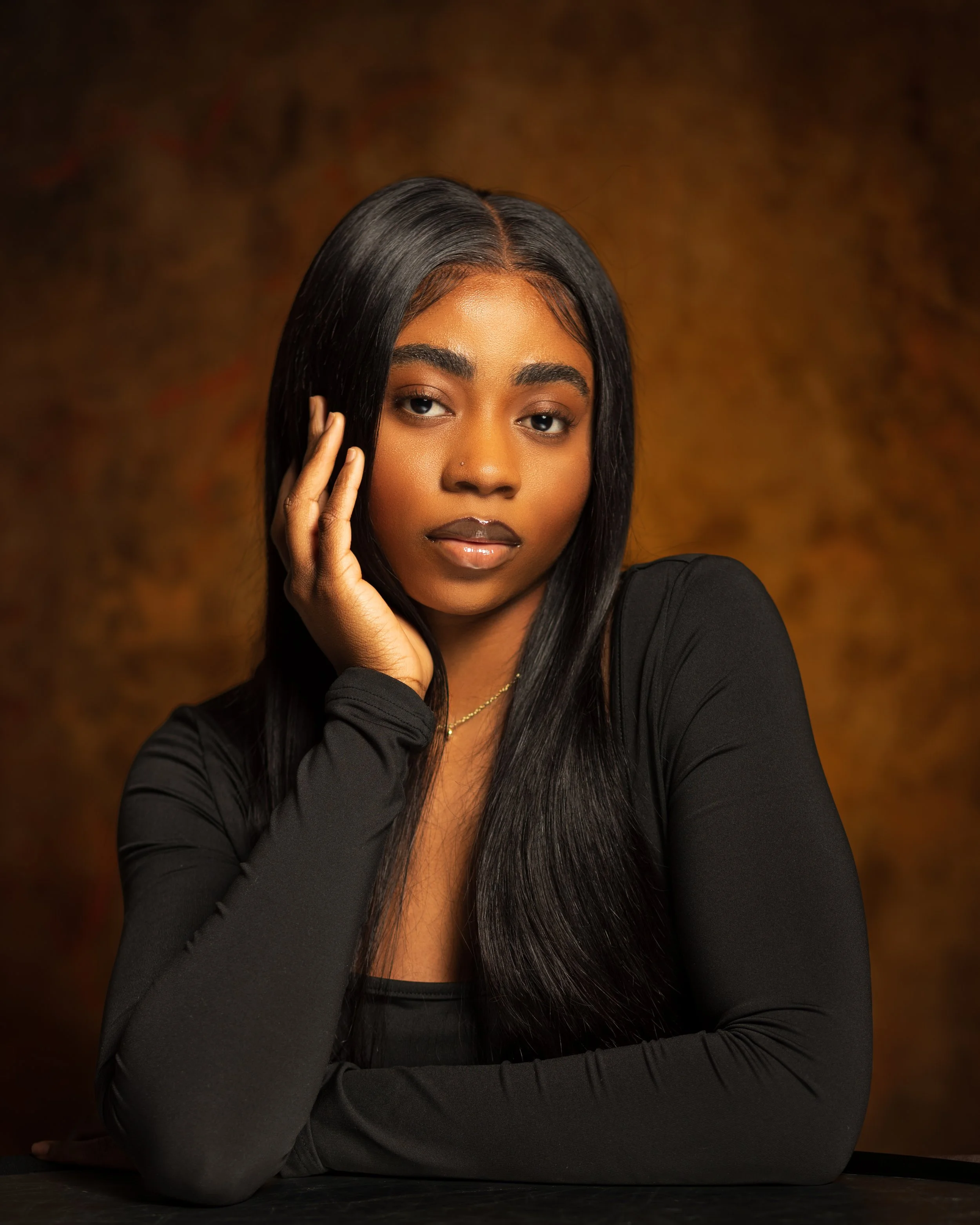 A young woman with long black hair, wearing a black long-sleeve top, resting her face on her hand, in front of a warm, textured background.