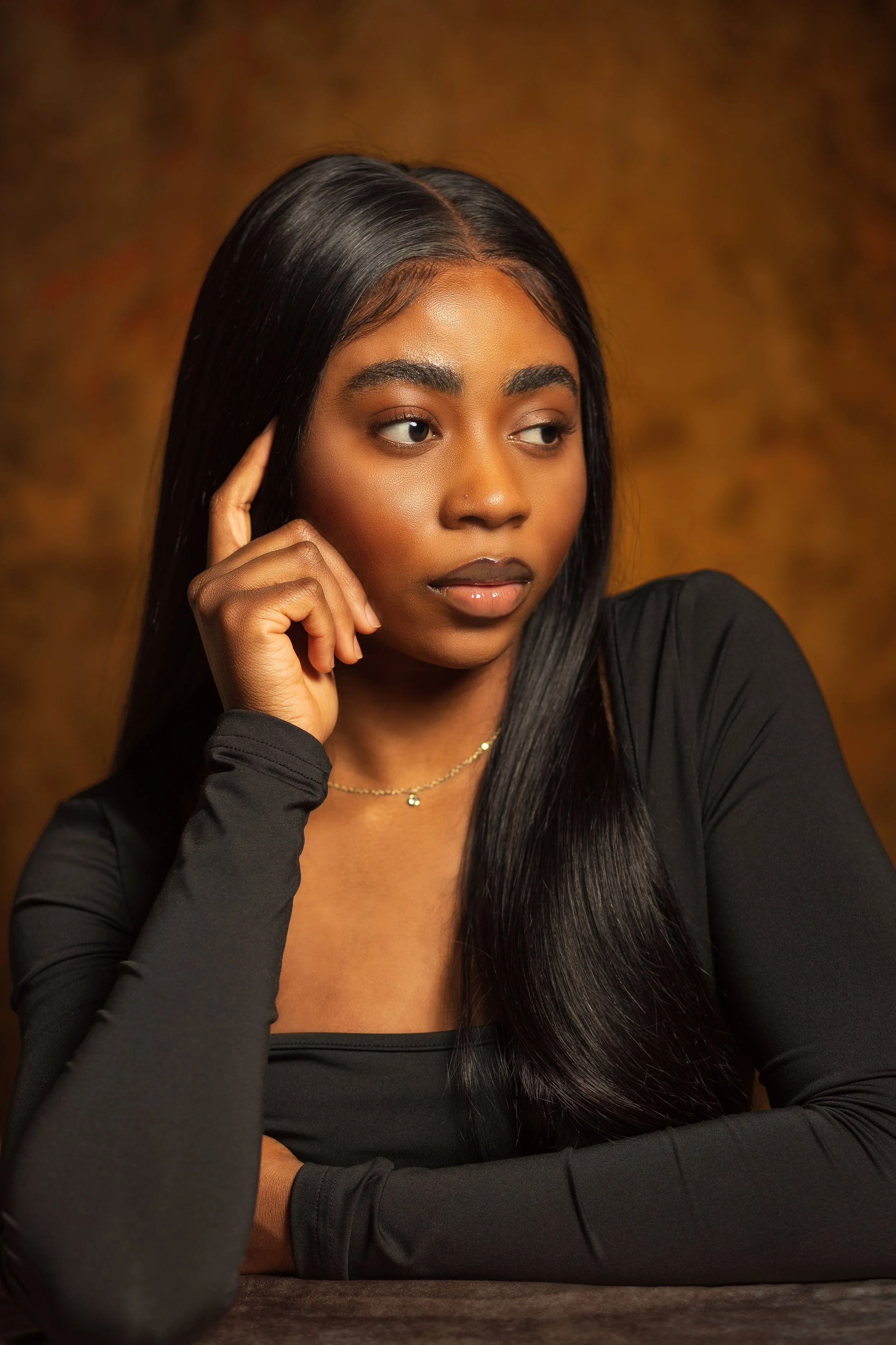 A woman with long black hair wearing a black top and a delicate necklace, resting her chin on her hand and looking to the side against a warm brown background.