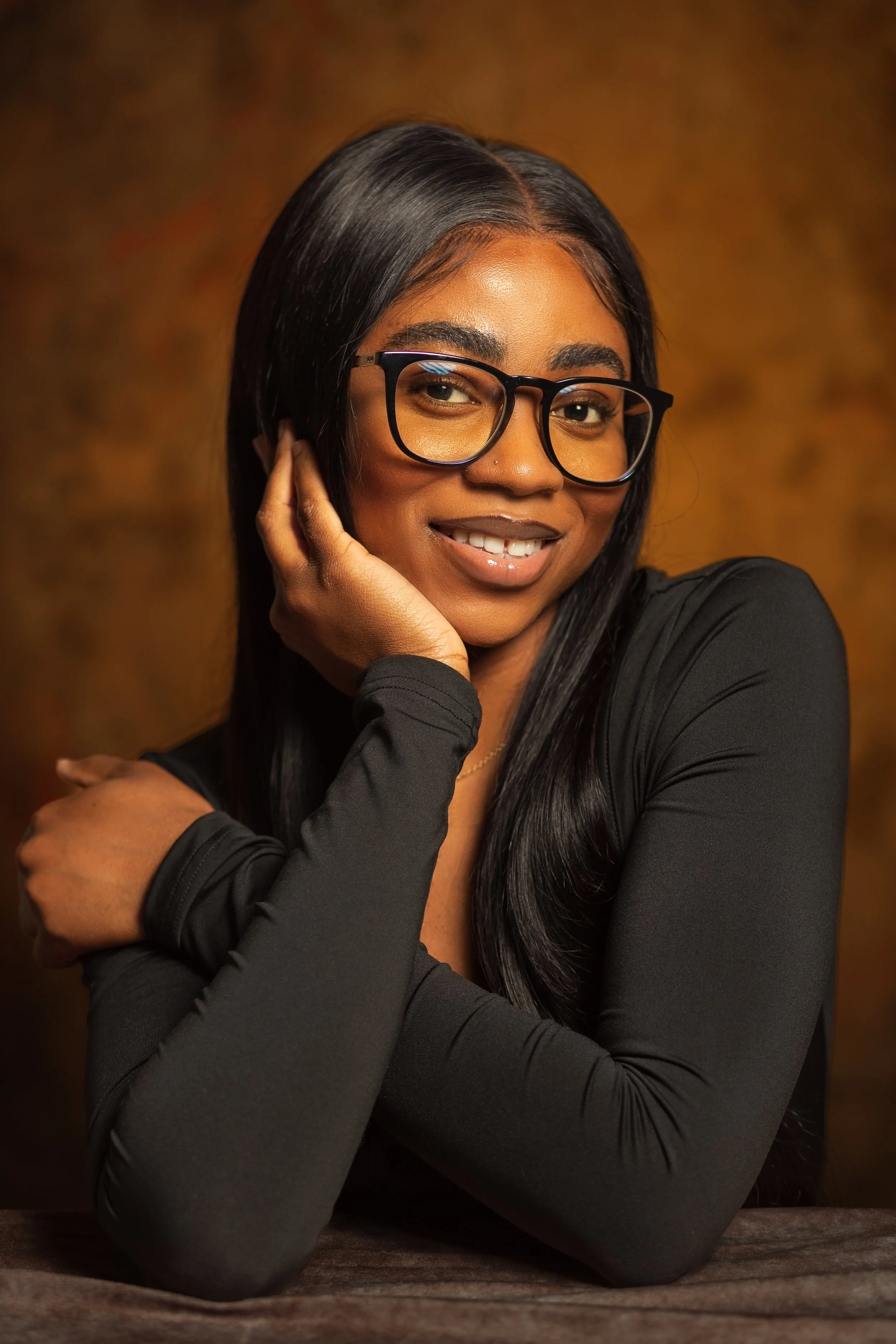 Portrait of a woman with long black hair, wearing glasses and a black top, smiling gently with her hand near her face, against a warm brown background.