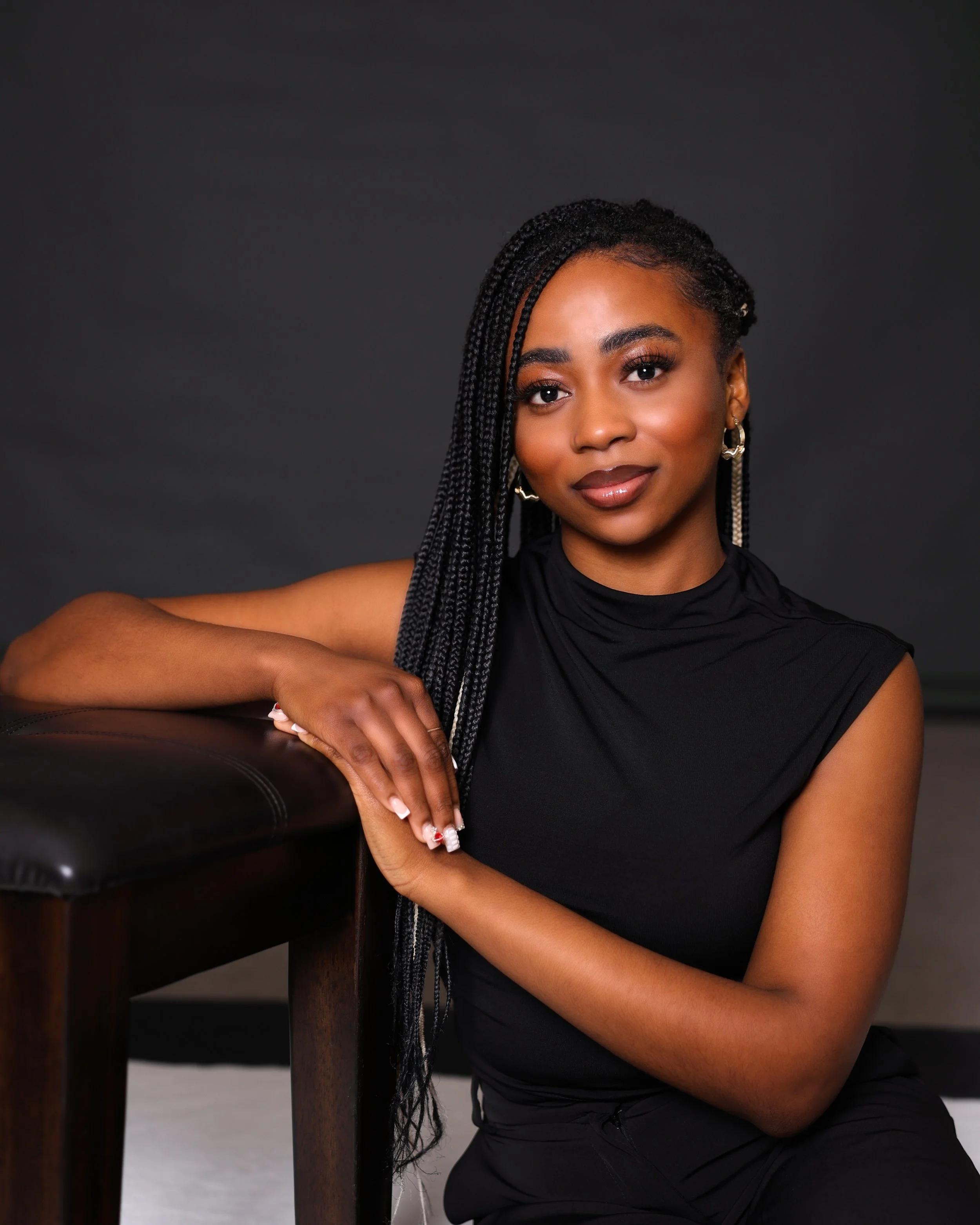 A woman with braided hair, wearing a black sleeveless top, sitting next to a dark wooden table with a leather top, in front of a dark background.