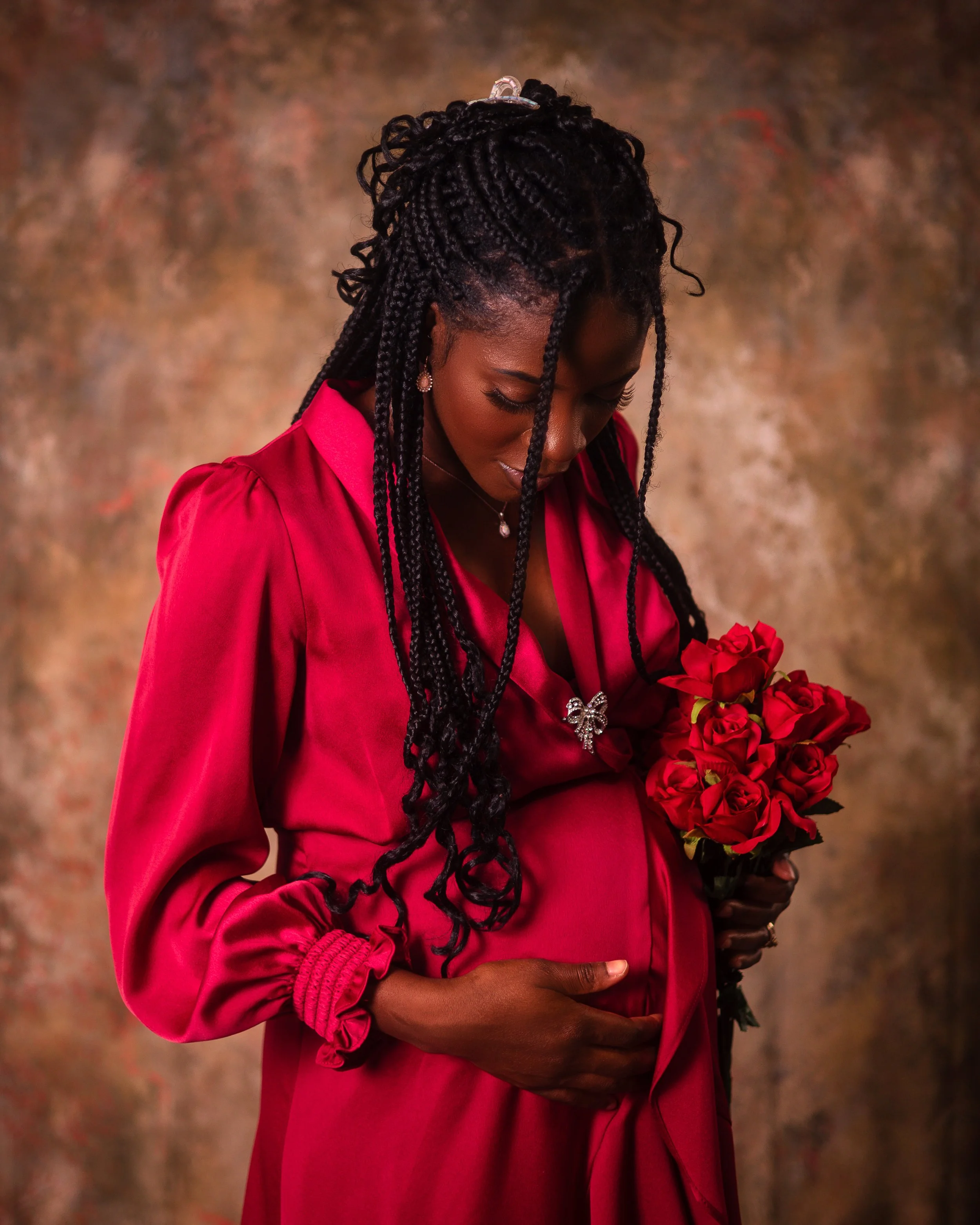 A pregnant woman dressed in a red gown holding a bouquet of red roses, looking down on her baby bump.
