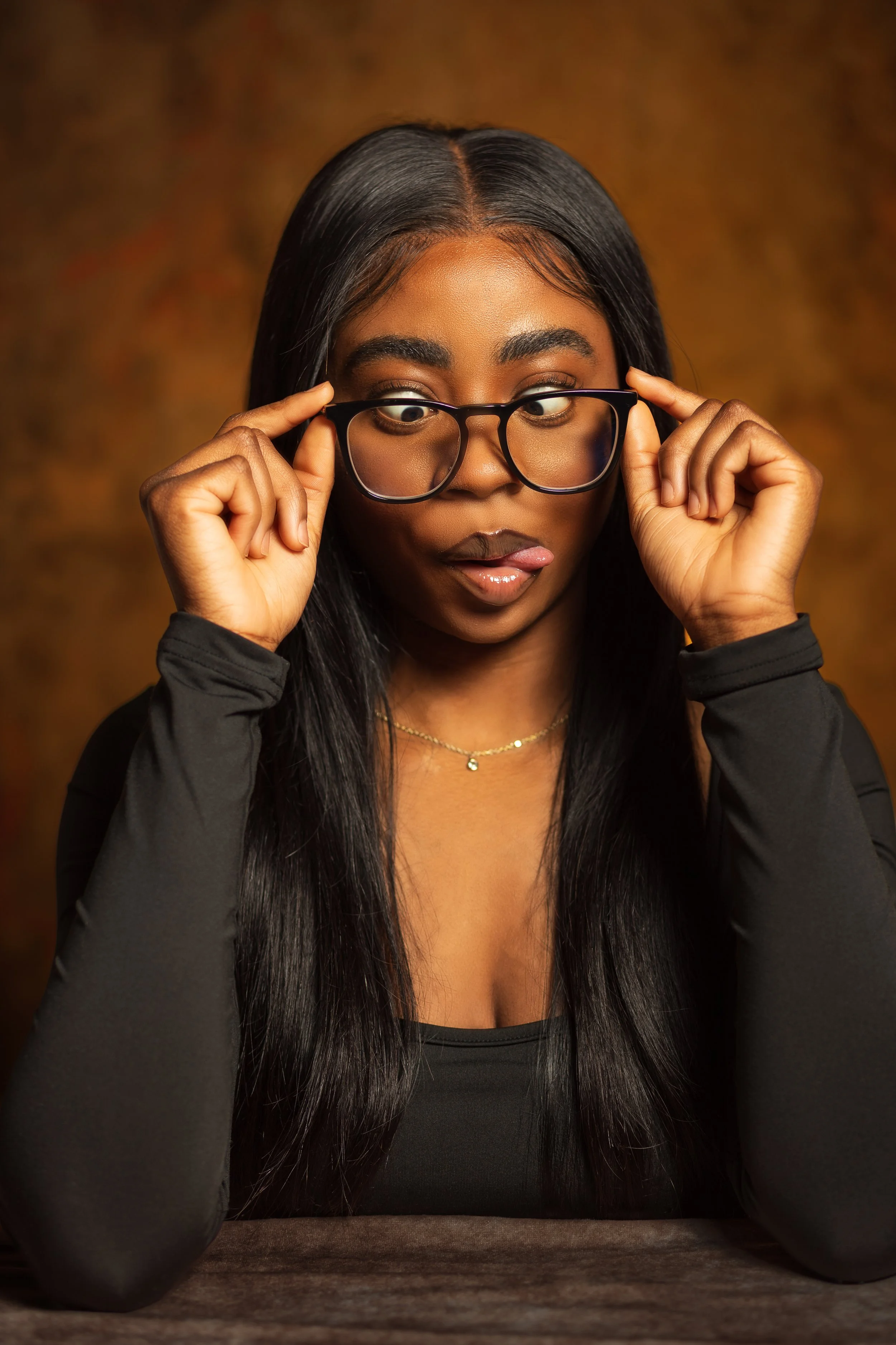 A young woman with long black hair and glasses pulling them down slightly and looking downwards, wearing a black top and a delicate necklace, seated at a table with a warm brown background.