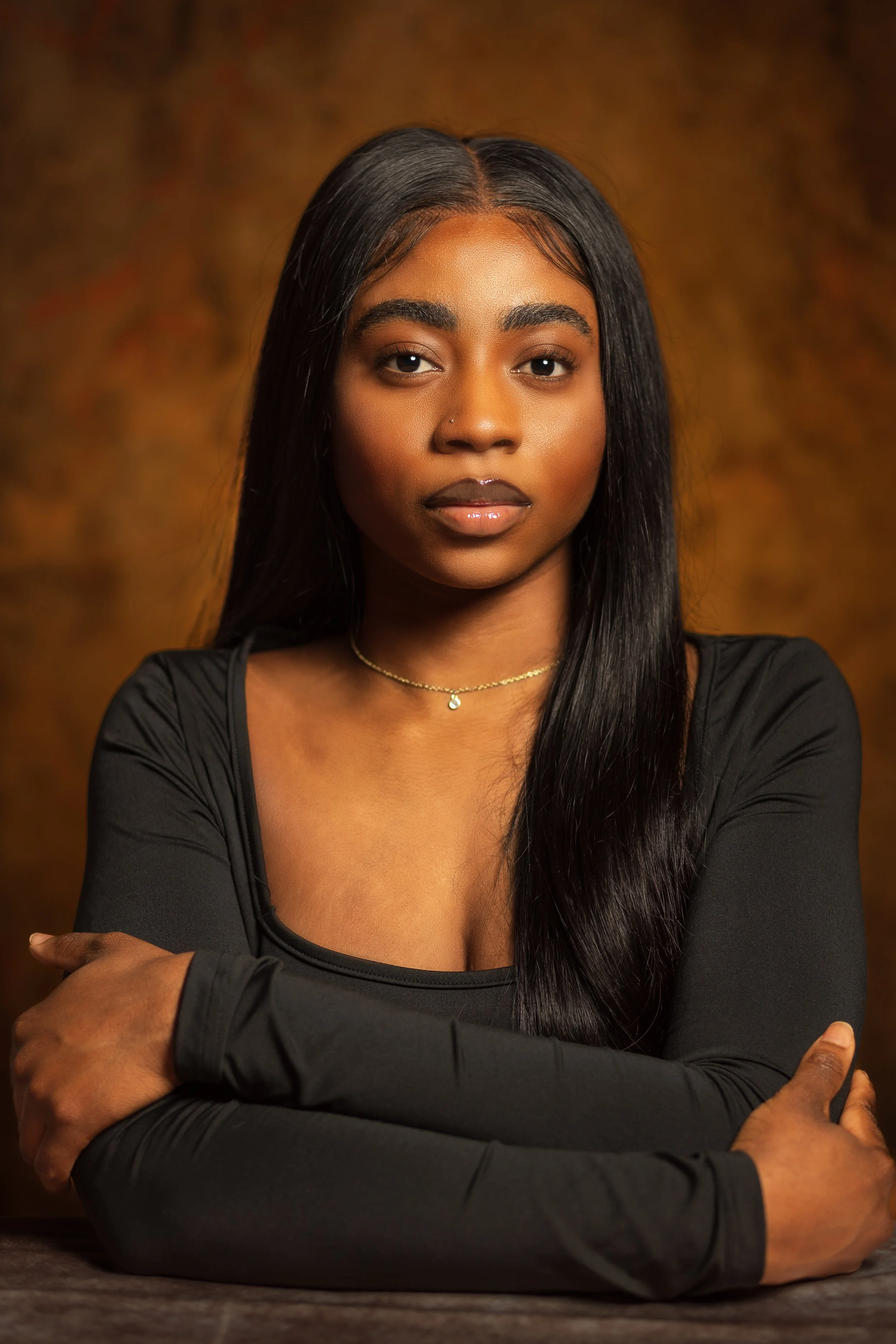 Portrait of a young Black woman with straight black hair, wearing a black top, a delicate necklace, and makeup, sitting in front of a warm, blurred background.