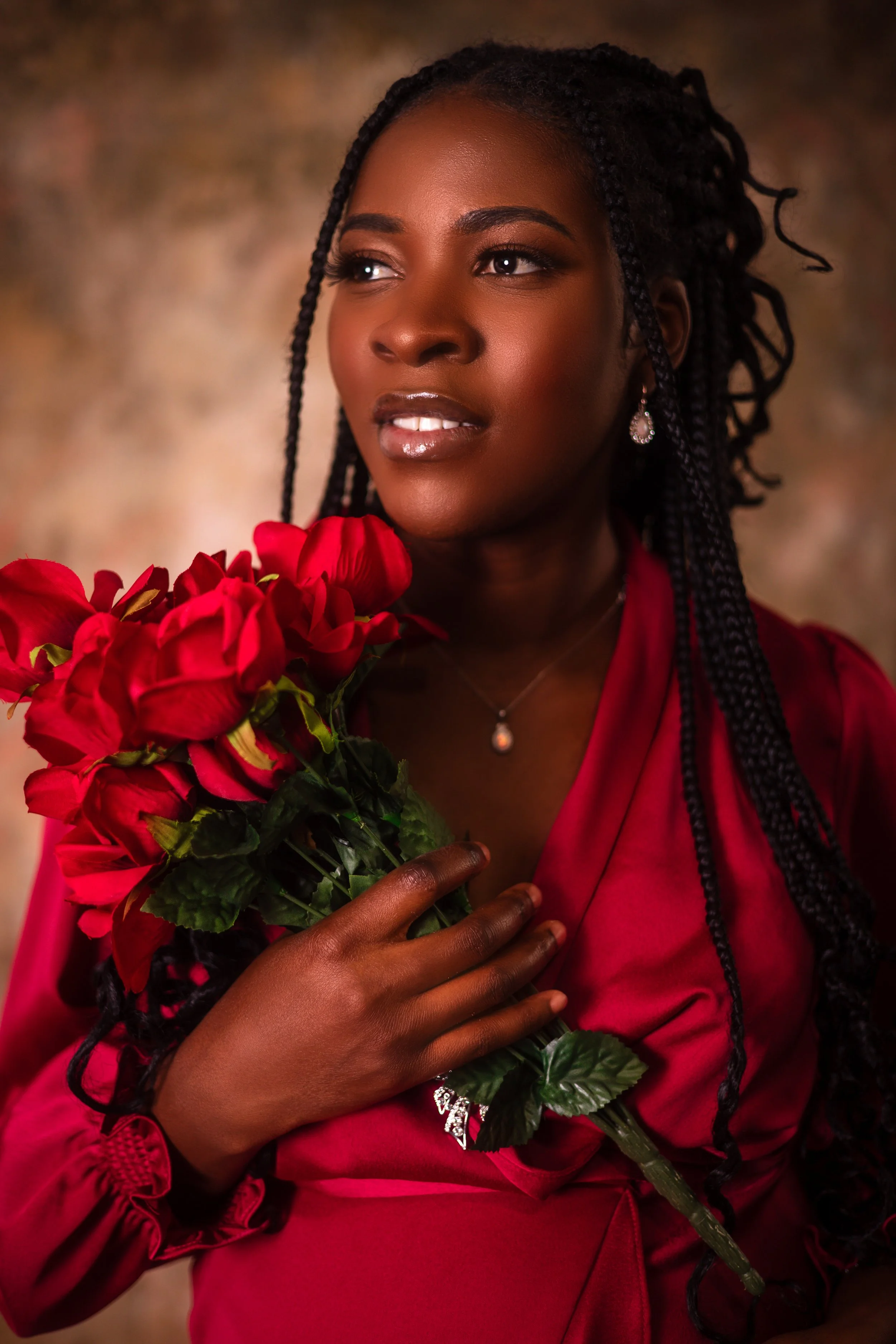 A woman with dark skin and braided hair is holding a bouquet of red flowers, wearing a red satin dress and jewelry, with a neutral background.