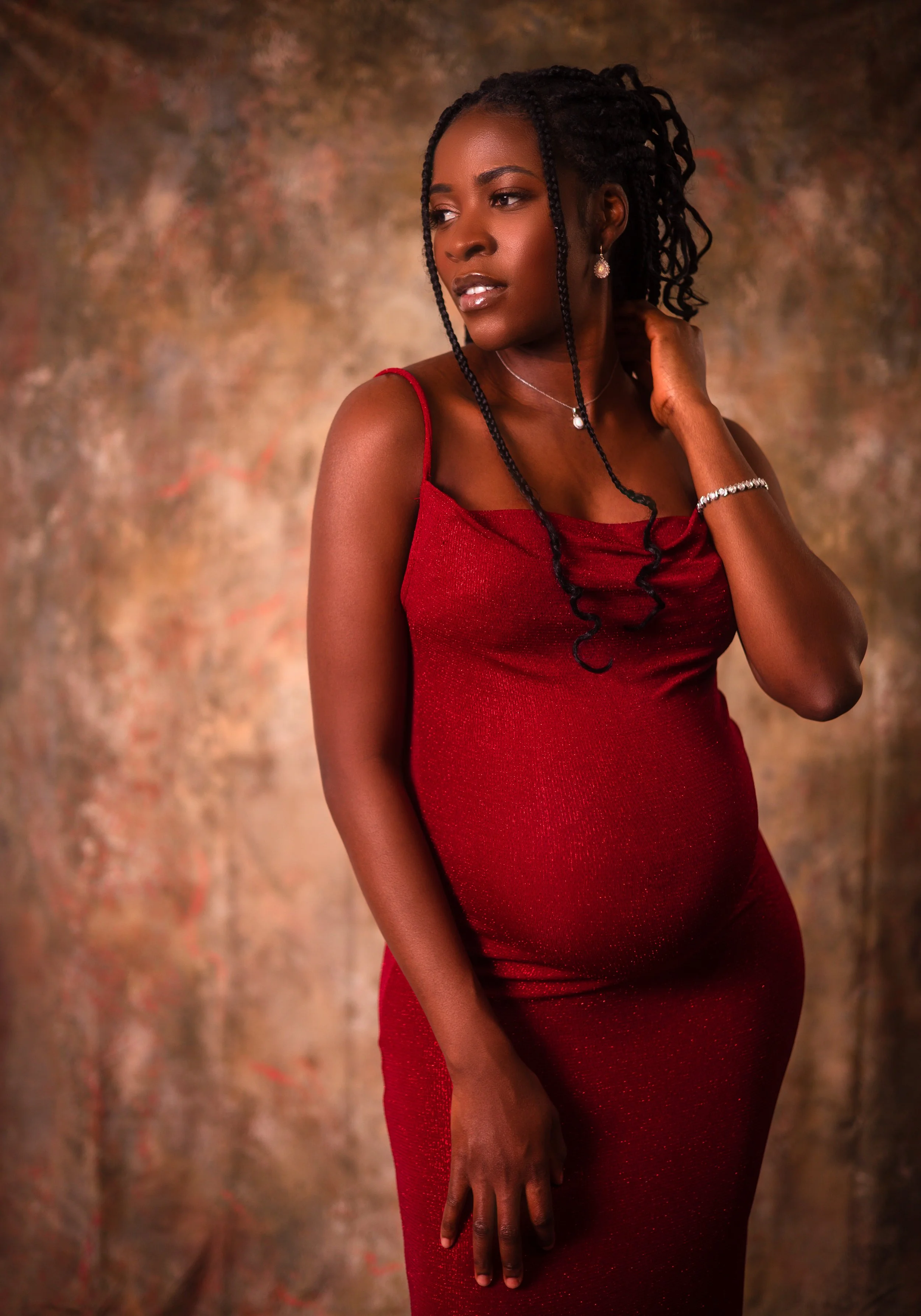 A pregnant woman with braided hair wearing a red, sleeveless dress and jewelry, standing against a textured, brownish background.
