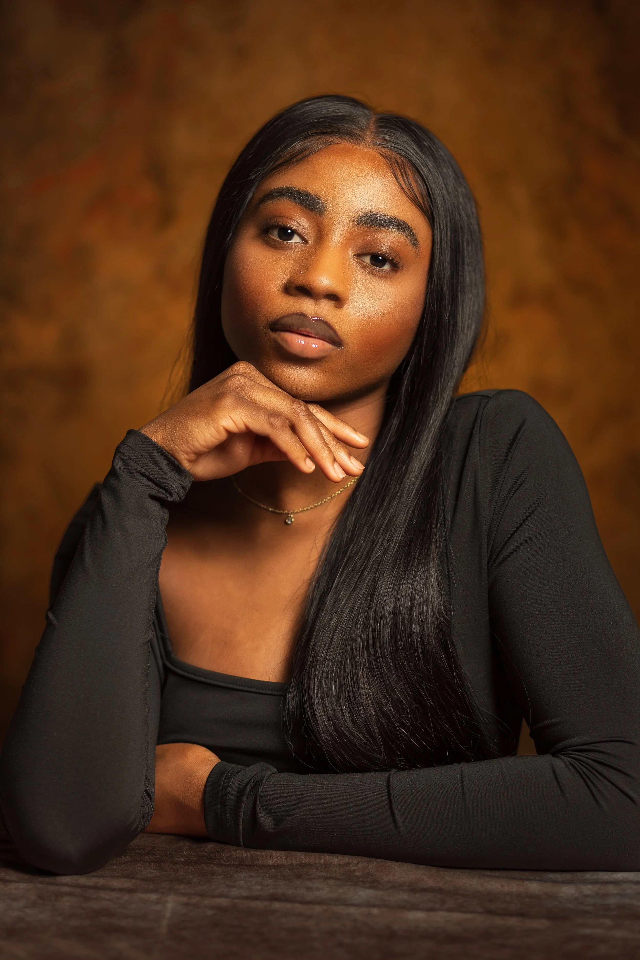 A young woman with long black hair, wearing a black top, resting her chin on her hand, with a warm blurred background.