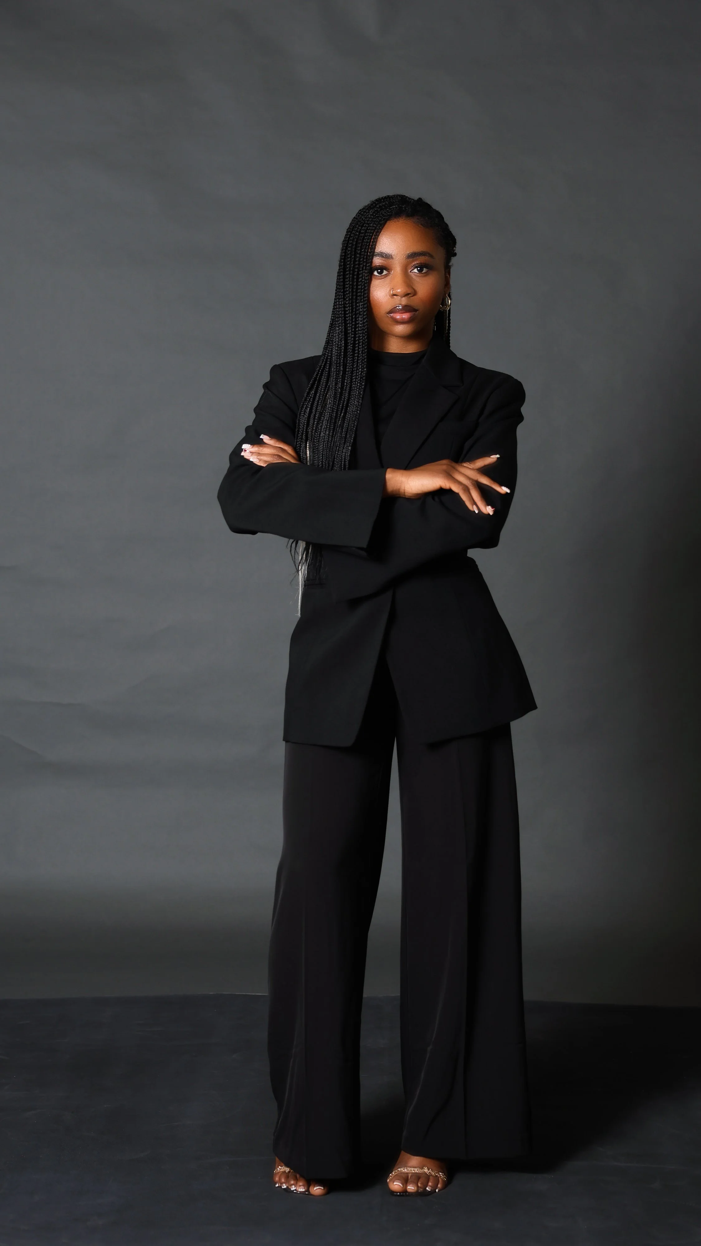 A woman with long braided hair standing with arms crossed wearing a black suit against a grey background.