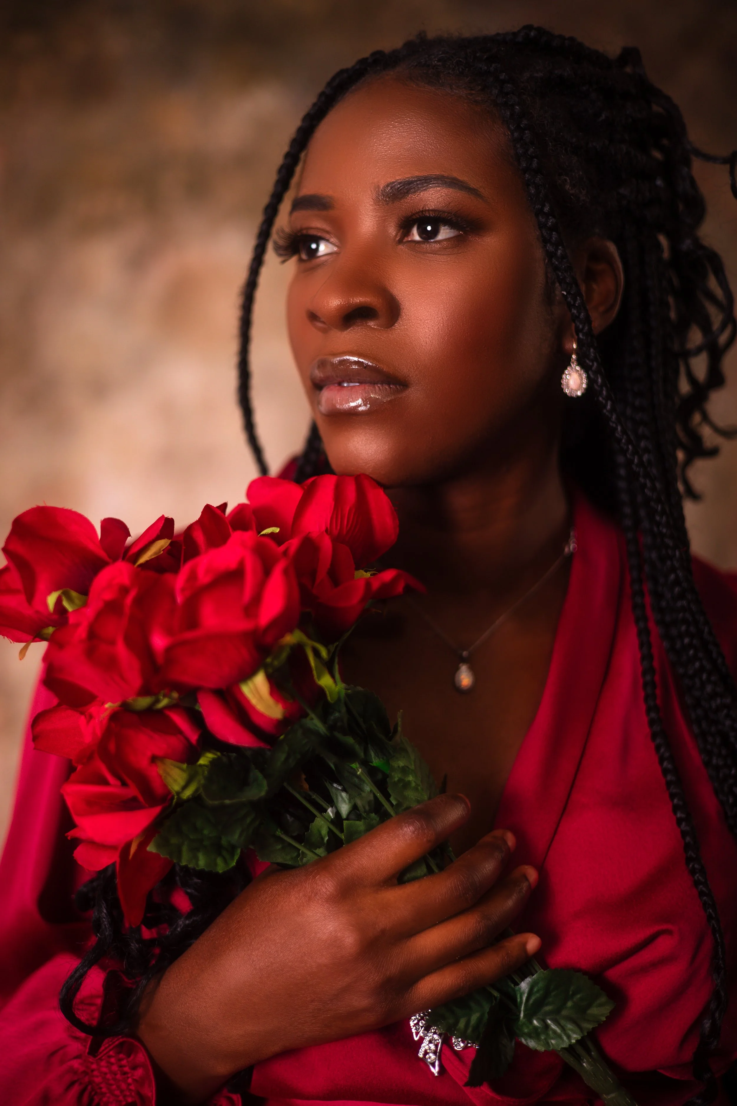 A woman with dark skin and long, braided hair holds a bouquet of red flowers close to her chest, wearing a deep red garment and elegant jewelry, with a thoughtful expression against a neutral background.
