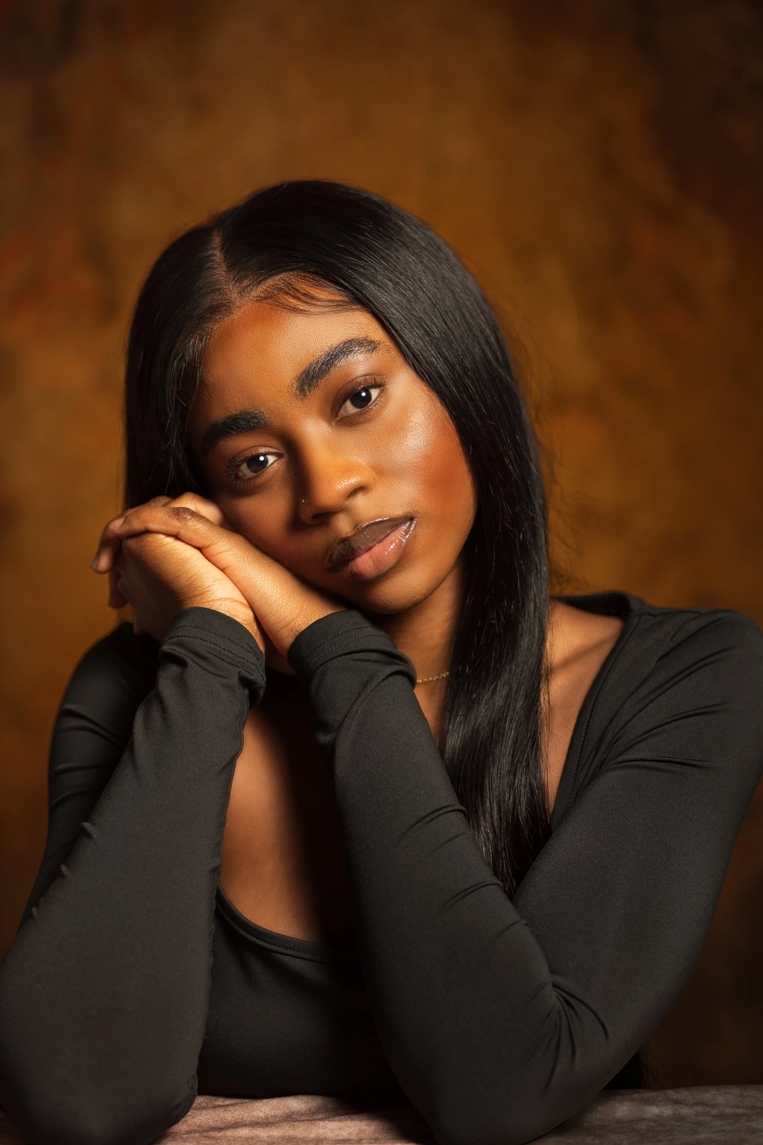 Portrait of a young woman with long black hair, resting her head on her hands, wearing a black long-sleeve top against a warm brown background.