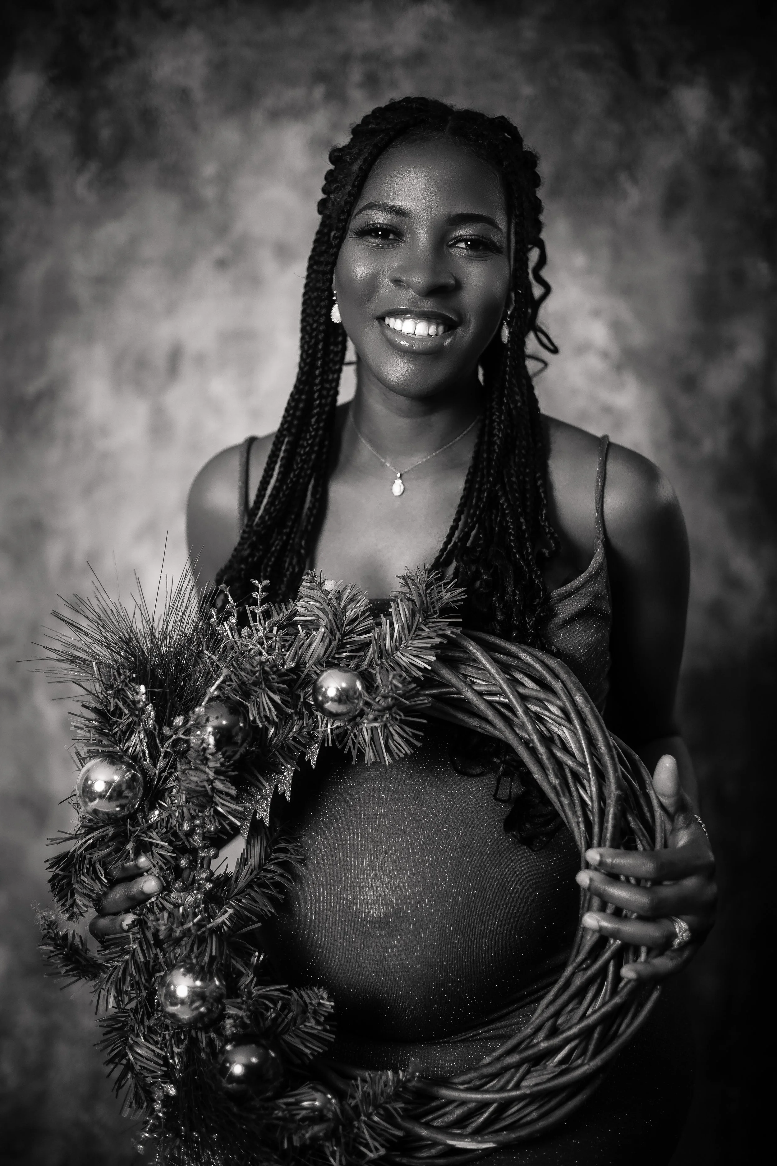 Black and white portrait of a smiling woman with braided hair, wearing a sleeveless dress, holding a Christmas wreath decorated with baubles and pine branches.