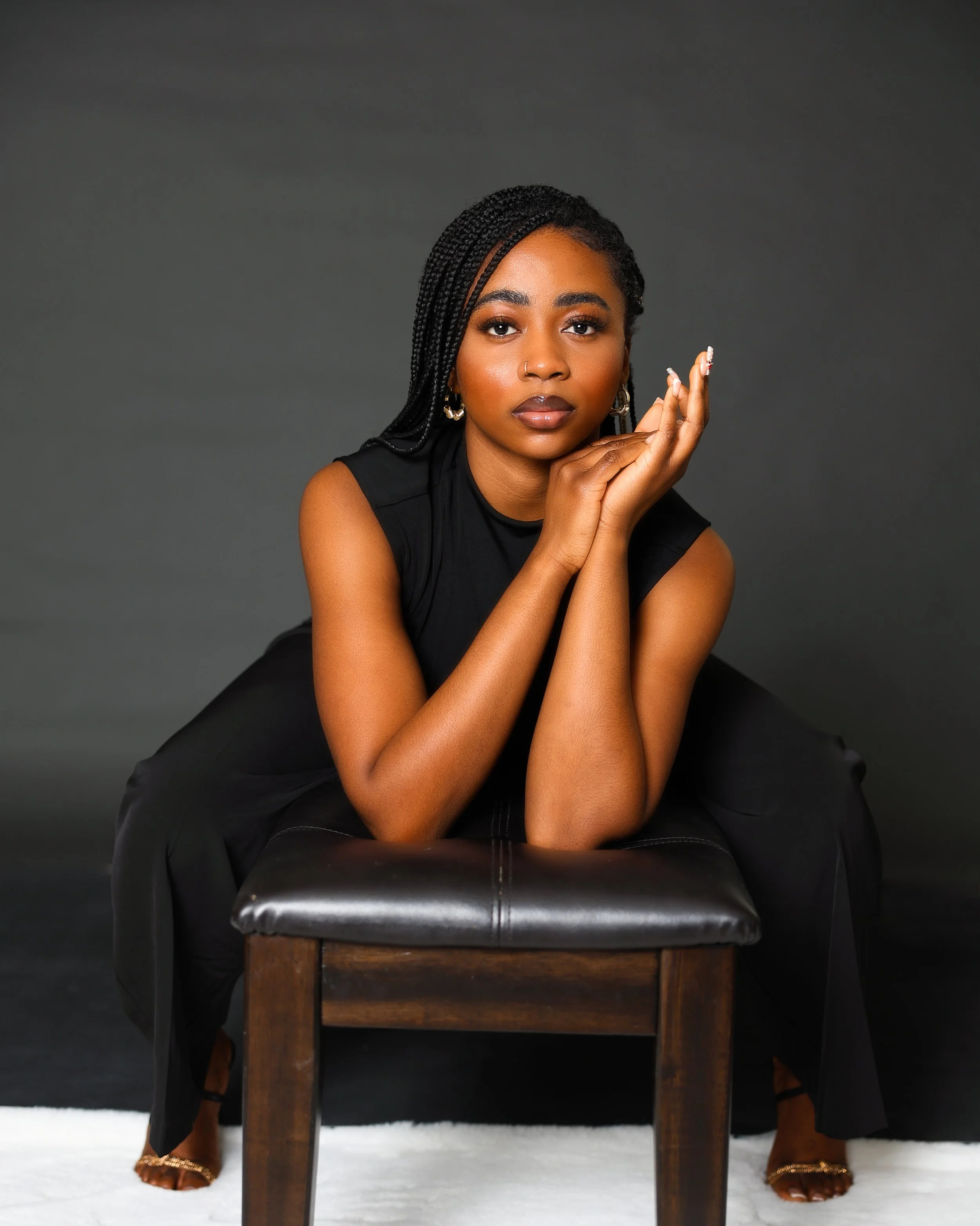 A young woman with braided hair and gold hoop earrings sitting on a black leather stool against a dark background, leaning forward with her chin resting on her hand, and looking directly at the camera.