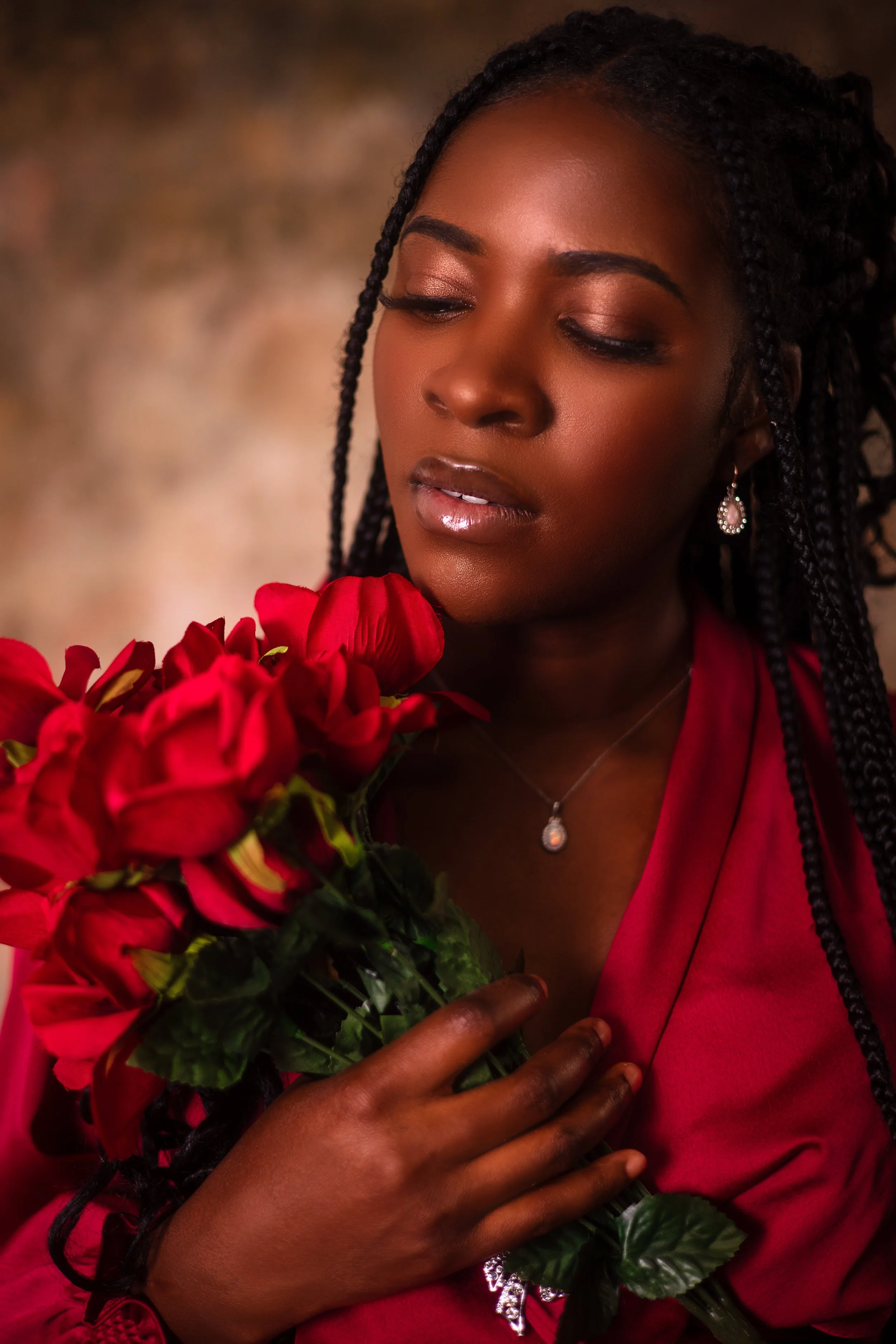 A woman with dark skin and long braided hair holding a bouquet of red flowers, wearing a red dress and jewelry, with a soft and warm background.