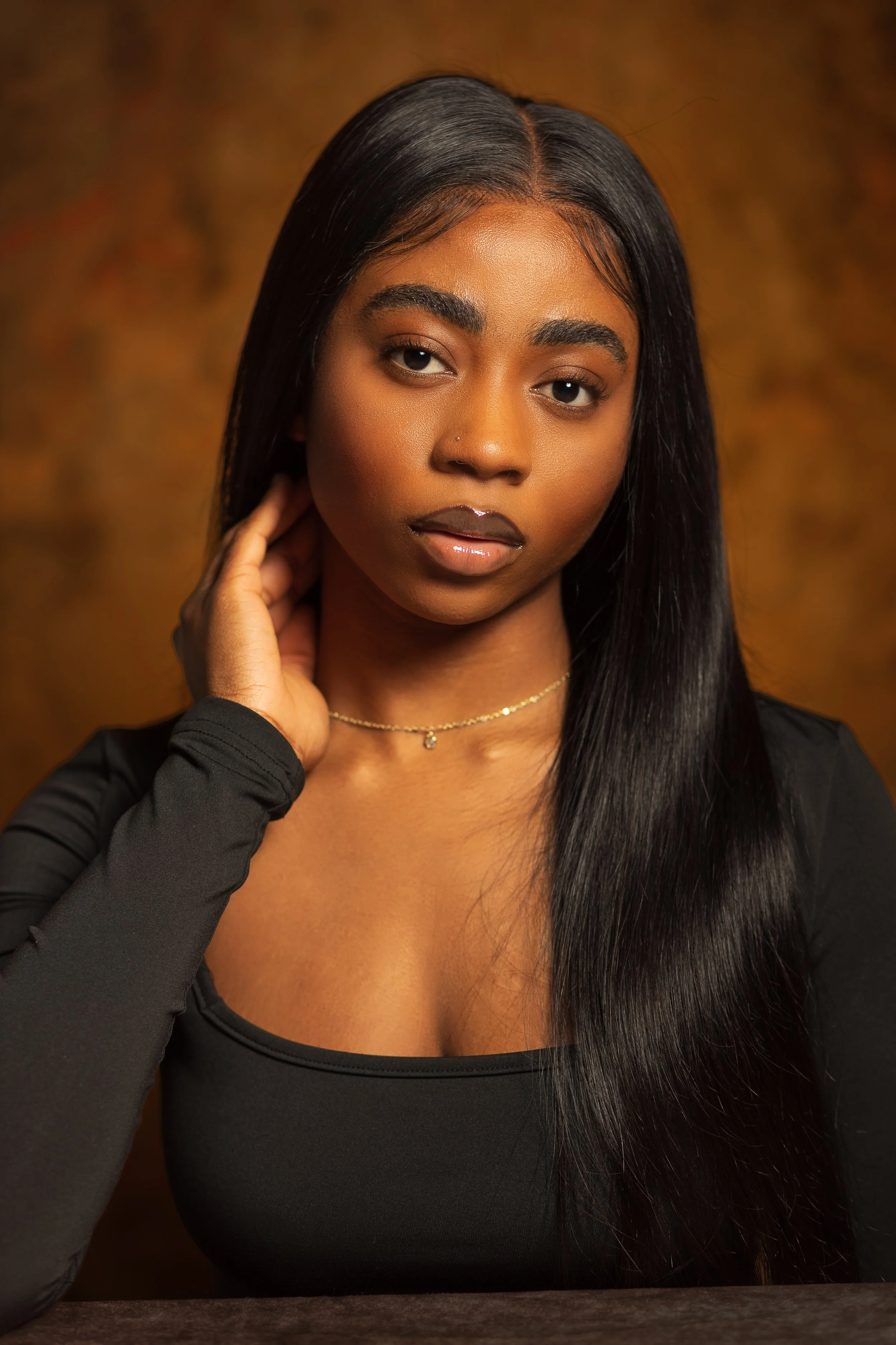 A young woman with long, straight black hair, wearing a black top and a delicate gold necklace, poses with her hand touching her neck against a warm brown background.