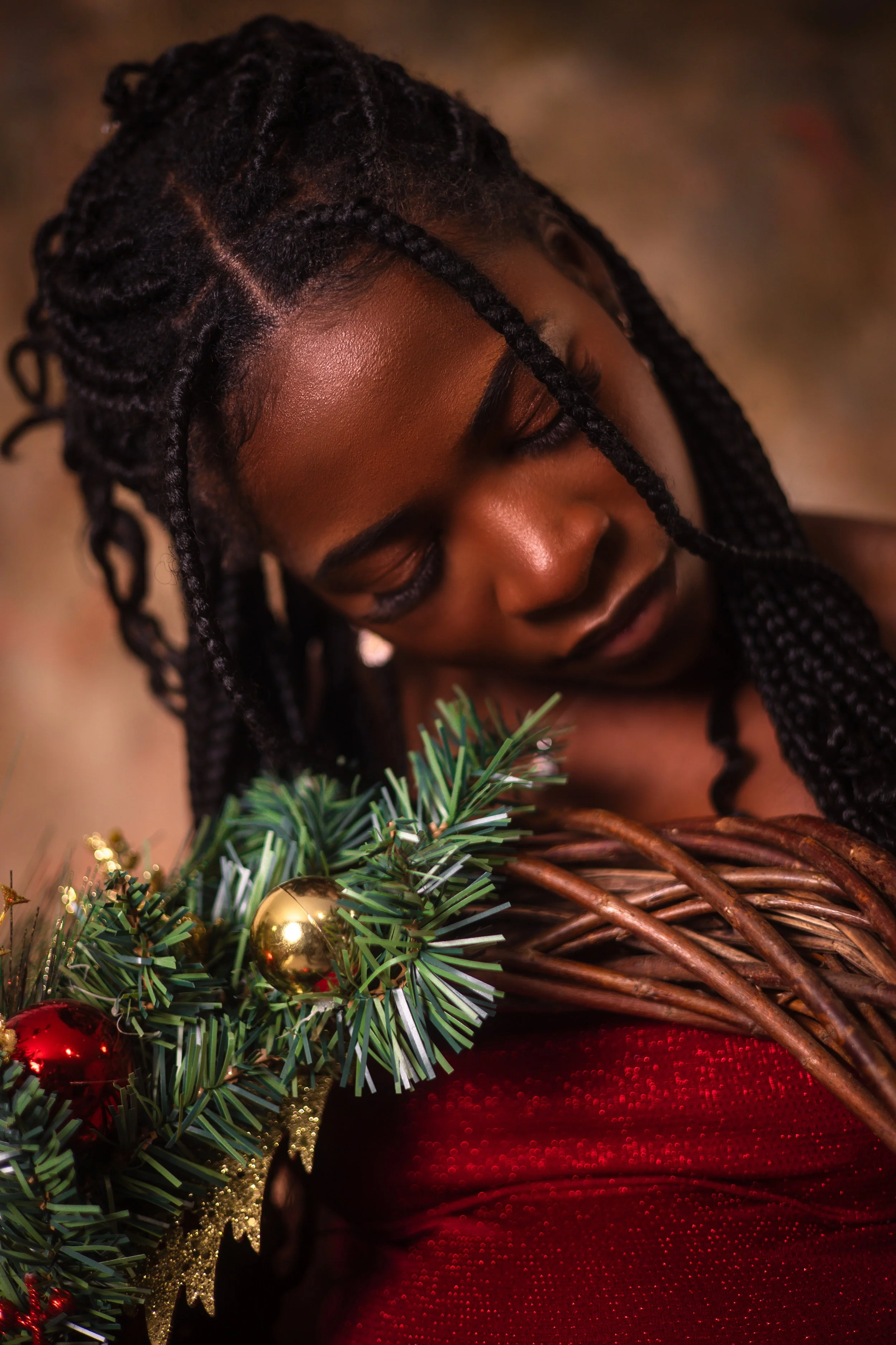 A woman with braided hair looking down at a Christmas wreath with ornaments, including a gold and a red bauble. She is wearing a red dress and has a serene expression.