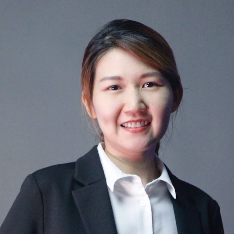 A woman with short brown hair wearing a black blazer and white shirt, smiling against a plain gray background.