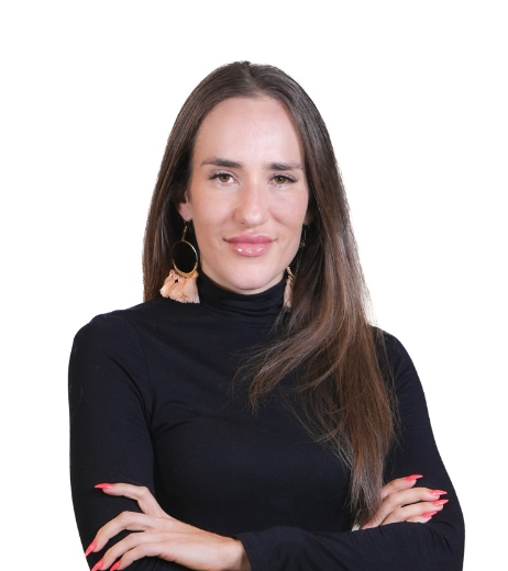 A woman with long brown hair, wearing a black turtleneck and large earrings, standing with arms crossed against a white background.