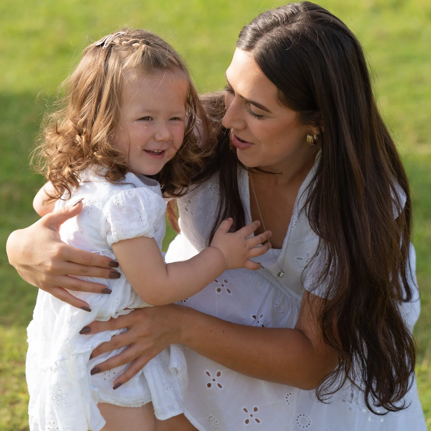 A woman and a young girl sharing a laugh outdoors on a grassy field, both dressed in white.