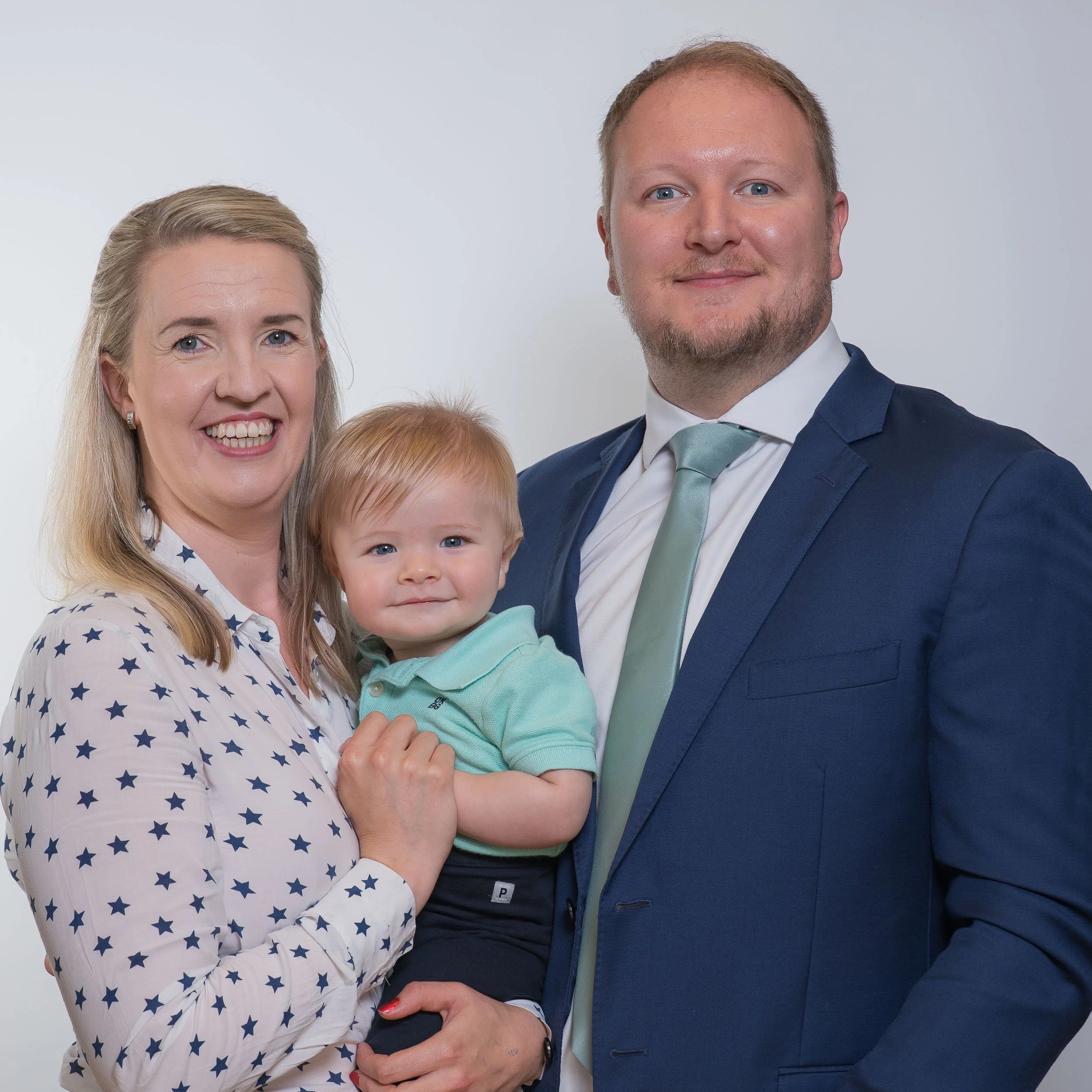 A family of three: mother, father, and a young child, posing together against a light background.