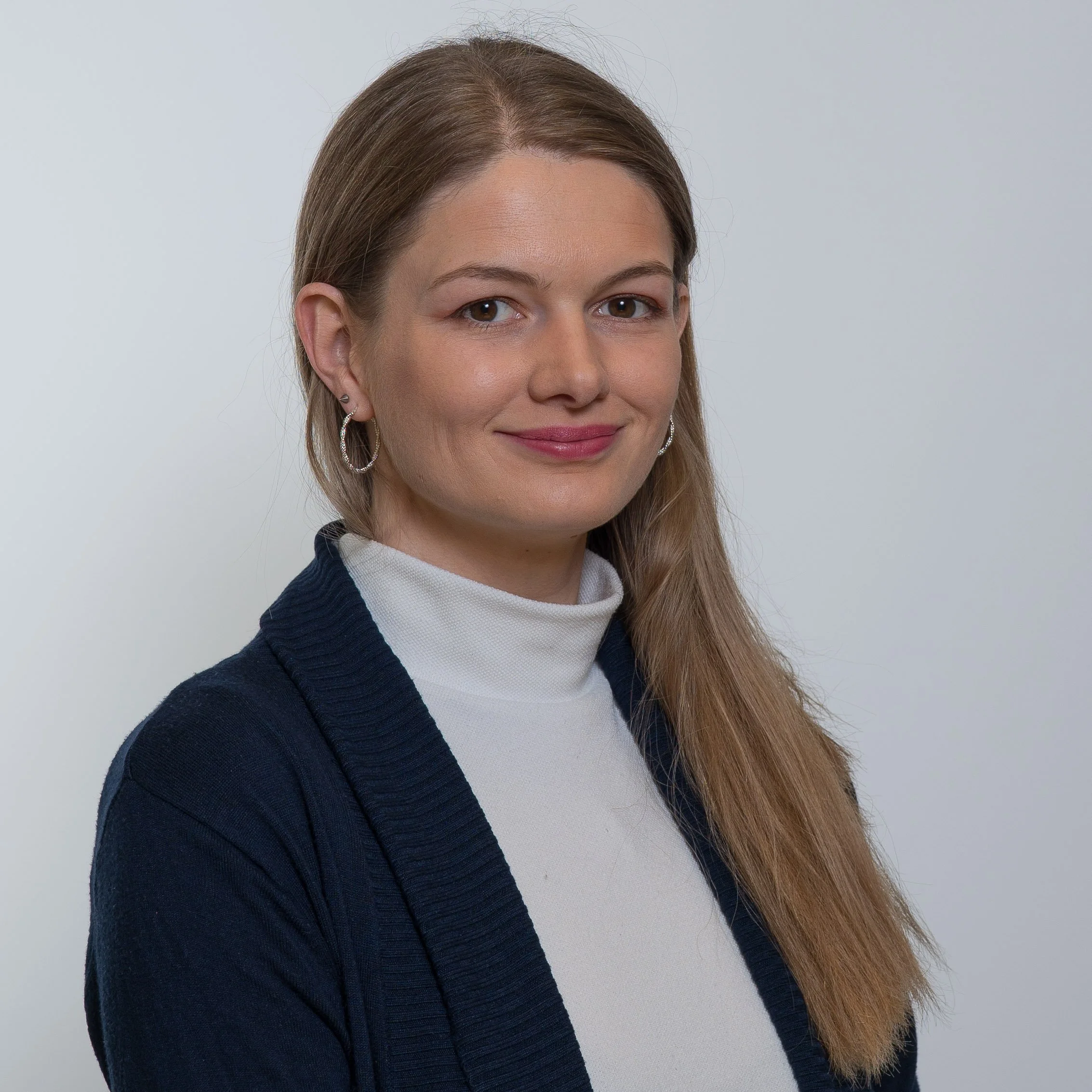 Portrait of a young woman with long light brown hair wearing a white turtleneck and navy cardigan, smiling with a plain light gray background.